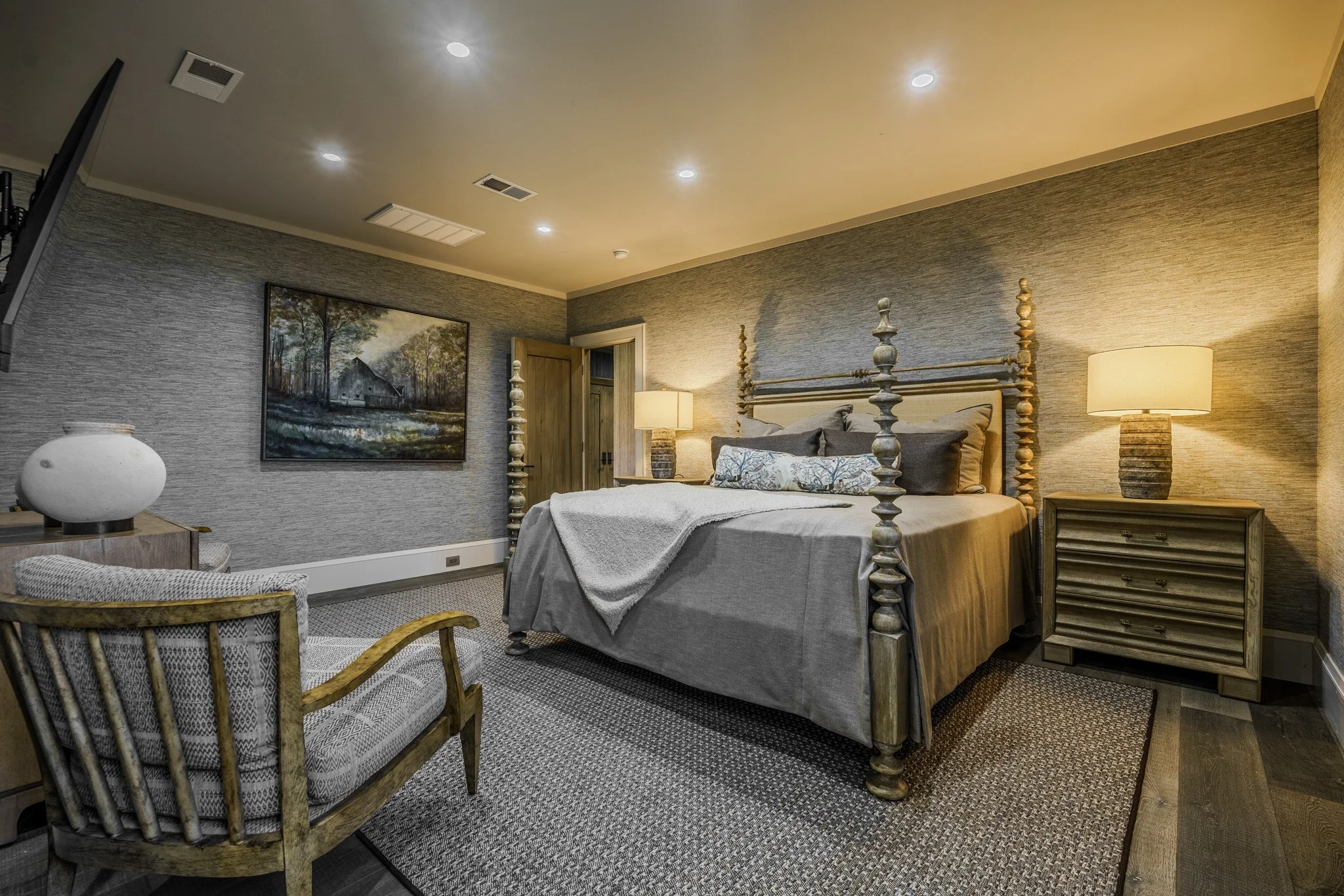 A cozy bedroom with a four-poster bed, a wooden armchair, and a landscape painting on the wall. Two bedside tables with lamps, textured wall covering, and a carpeted floor.
