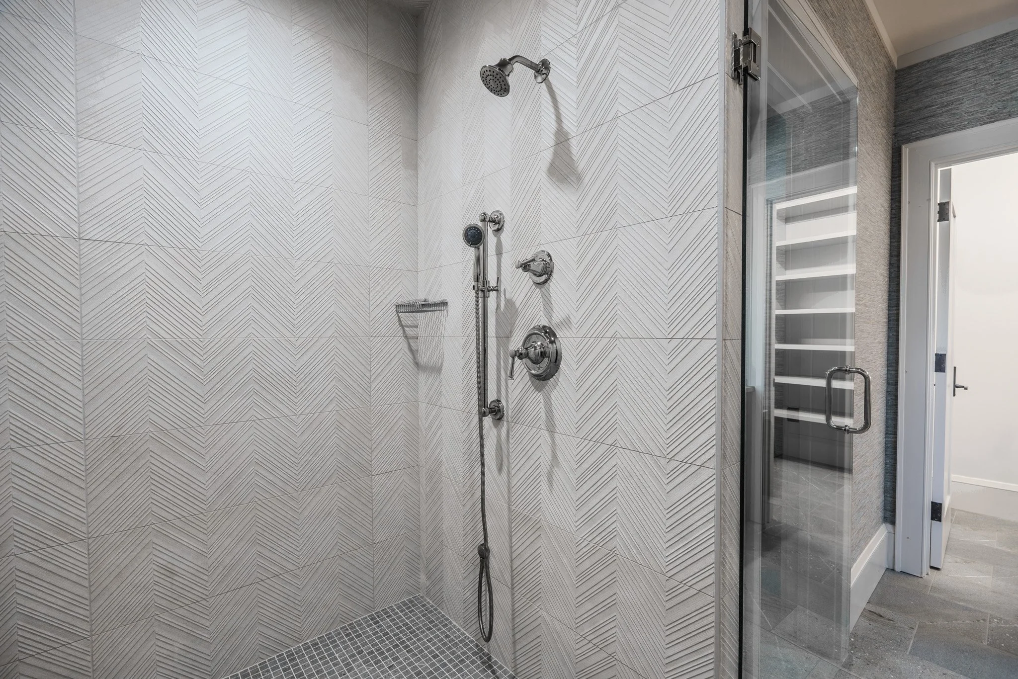Modern shower area with decorative tile walls, a handheld shower head, and a glass door leading to a closet with shelves.