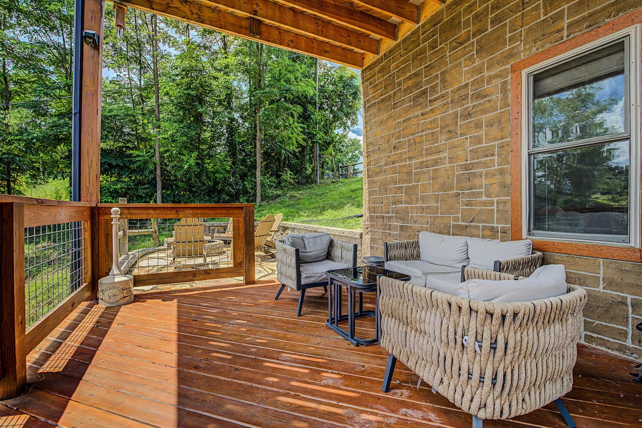 A wooden deck patio with outdoor furniture, including cushioned chairs and a firepit, attached to a house with a brick exterior wall and a window, surrounded by green trees and a grassy yard.