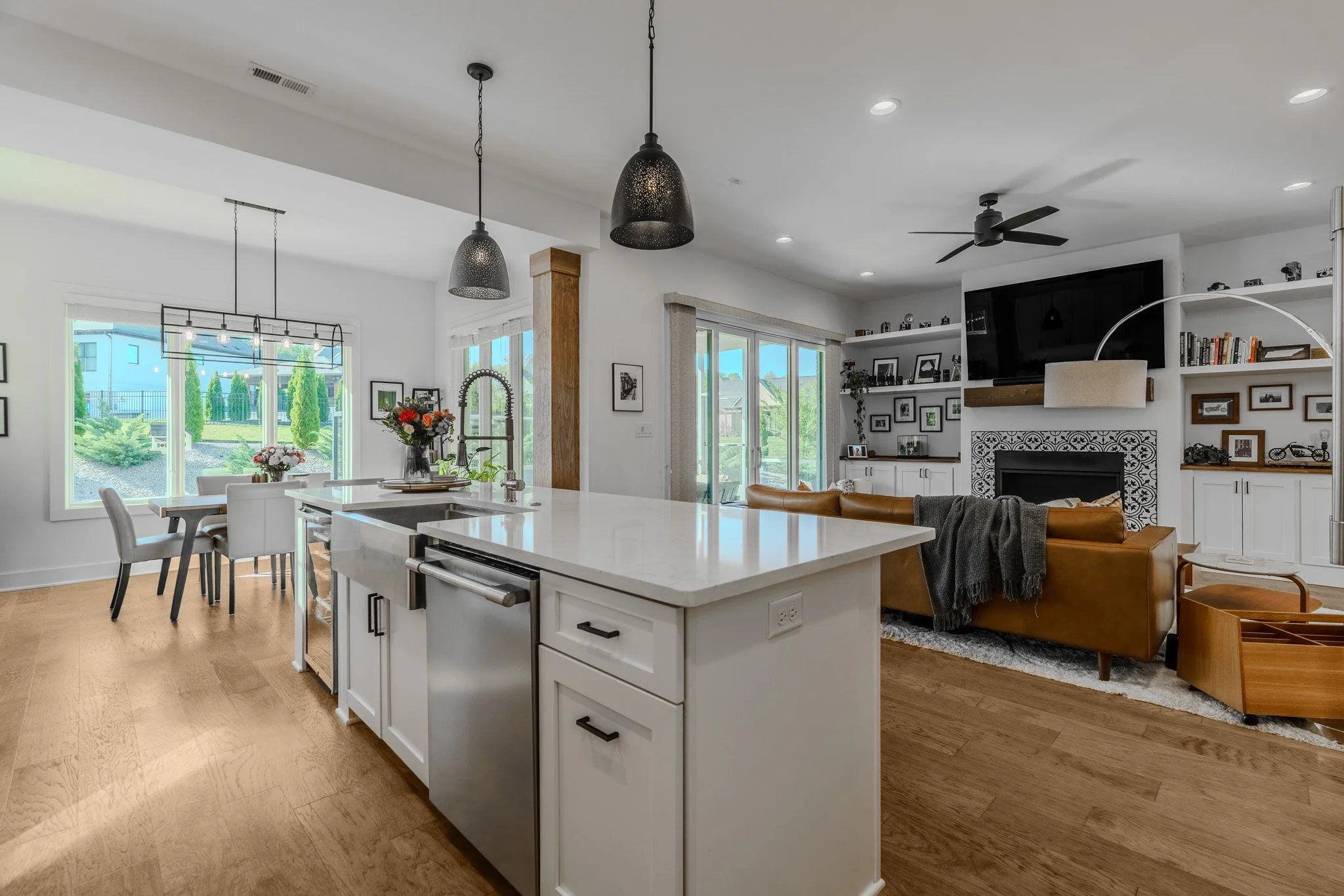 Open-concept kitchen and living room with white cabinetry, hardwood floors, a fireplace with black and white patterned tile, and large windows with outdoor views.