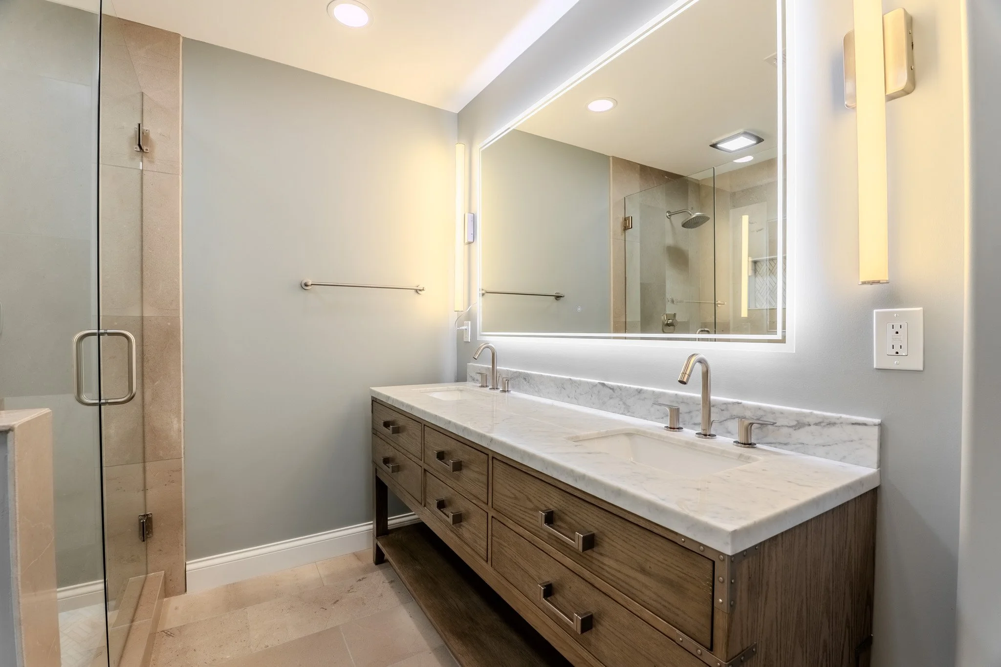 Modern bathroom with double sink marble countertop, large illuminated mirror, glass shower with walk-in design, beige tiled walls, and minimalist fixtures.