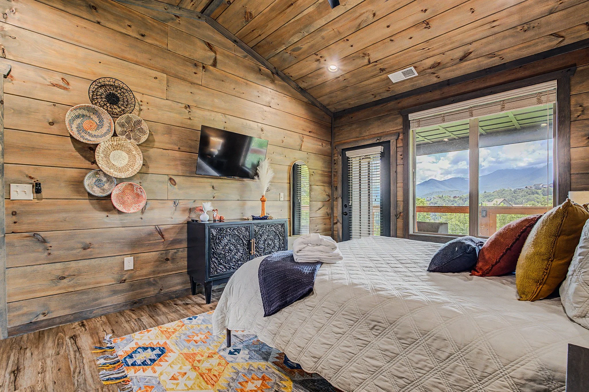 A bedroom with wooden walls and ceiling, a large window with a view of mountains, a bed with a white quilt and various pillows, a black ornate cabinet, and a colorful patterned rug.