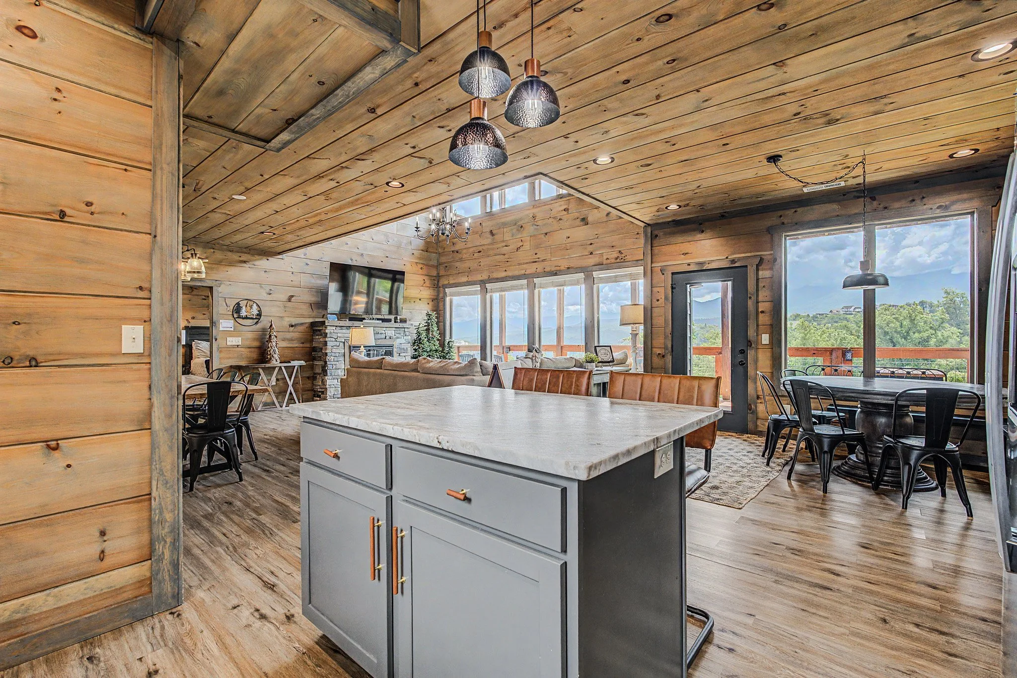 Interior of a rustic-style living space with a vaulted wooden ceiling, large windows showing greenery outside, a stone fireplace, a television, a chandelier, pendant lights, a dining table with chairs, and a kitchen island with a marble countertop.