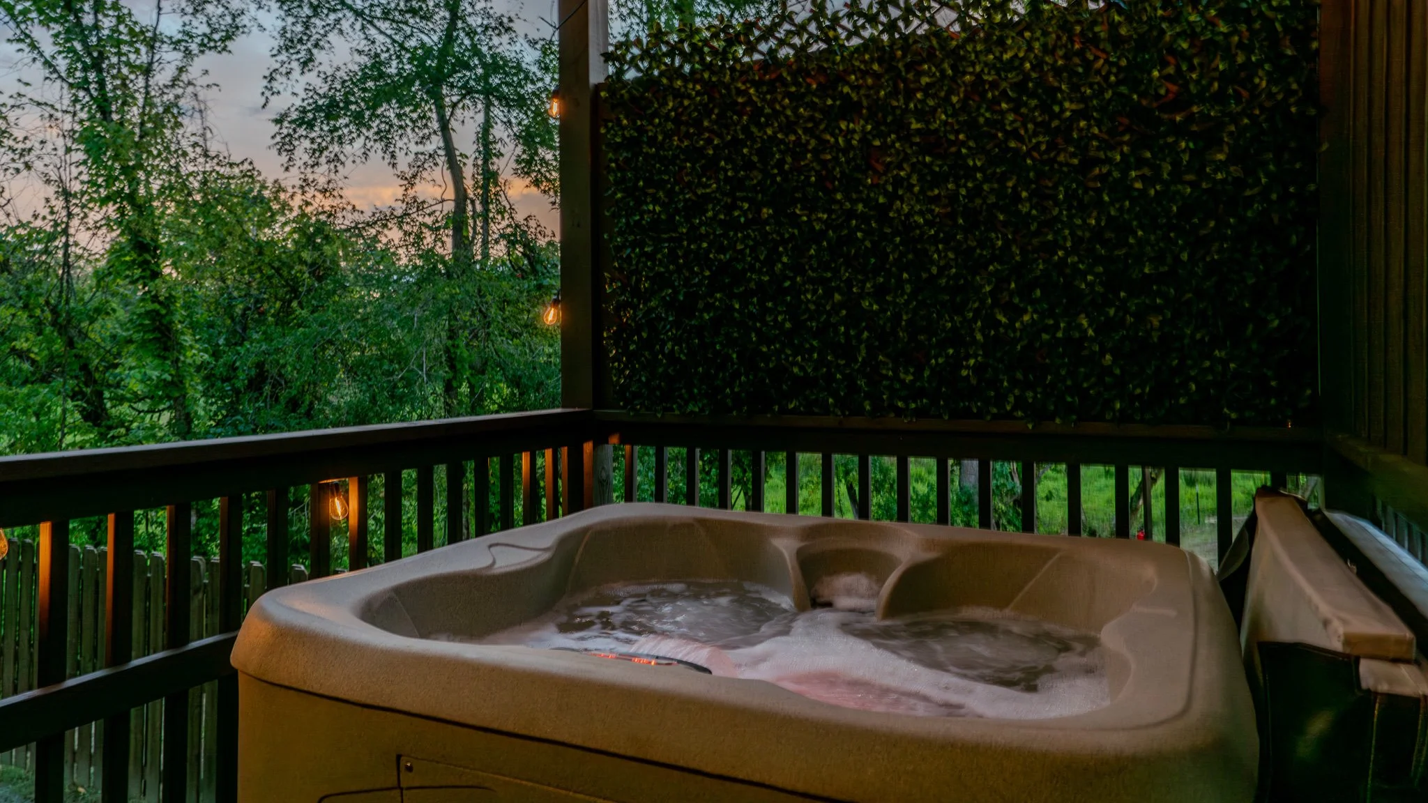 Outdoor hot tub on a balcony with greenery and trees in the background at dusk, decorated with string lights.