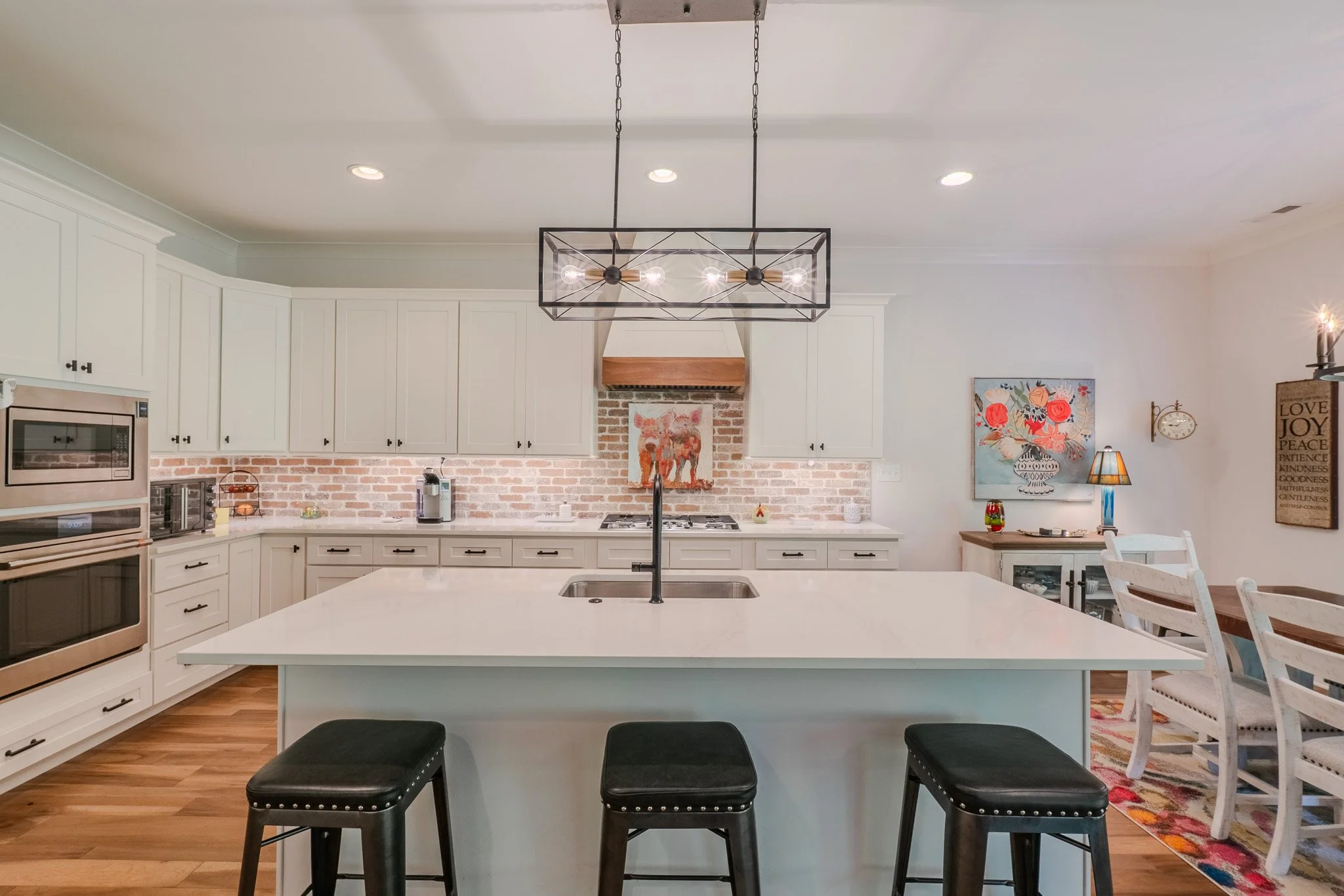 Modern kitchen with white cabinets, a large island with a white countertop, black stools, an exposed brick backsplash, and a dining area with white chairs and artwork on the wall.