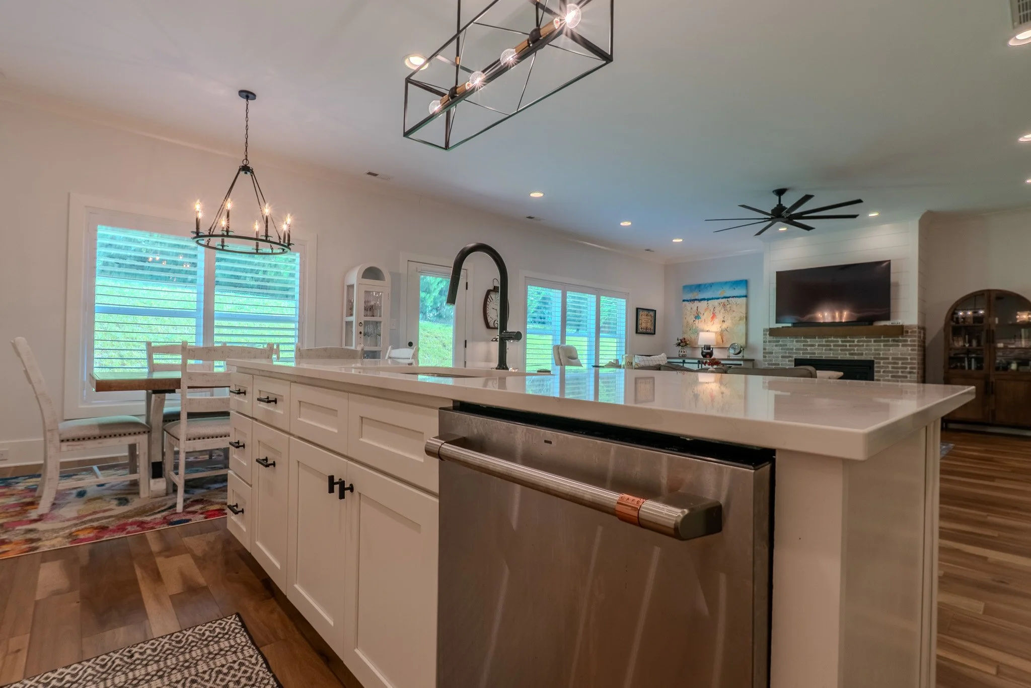 Modern open-concept kitchen with white cabinets, a large island, stainless steel dishwasher, black faucet, and a dining area with white chairs and a colorful rug. The background features a living space with a fireplace, flat-screen TV, artwork, and l
