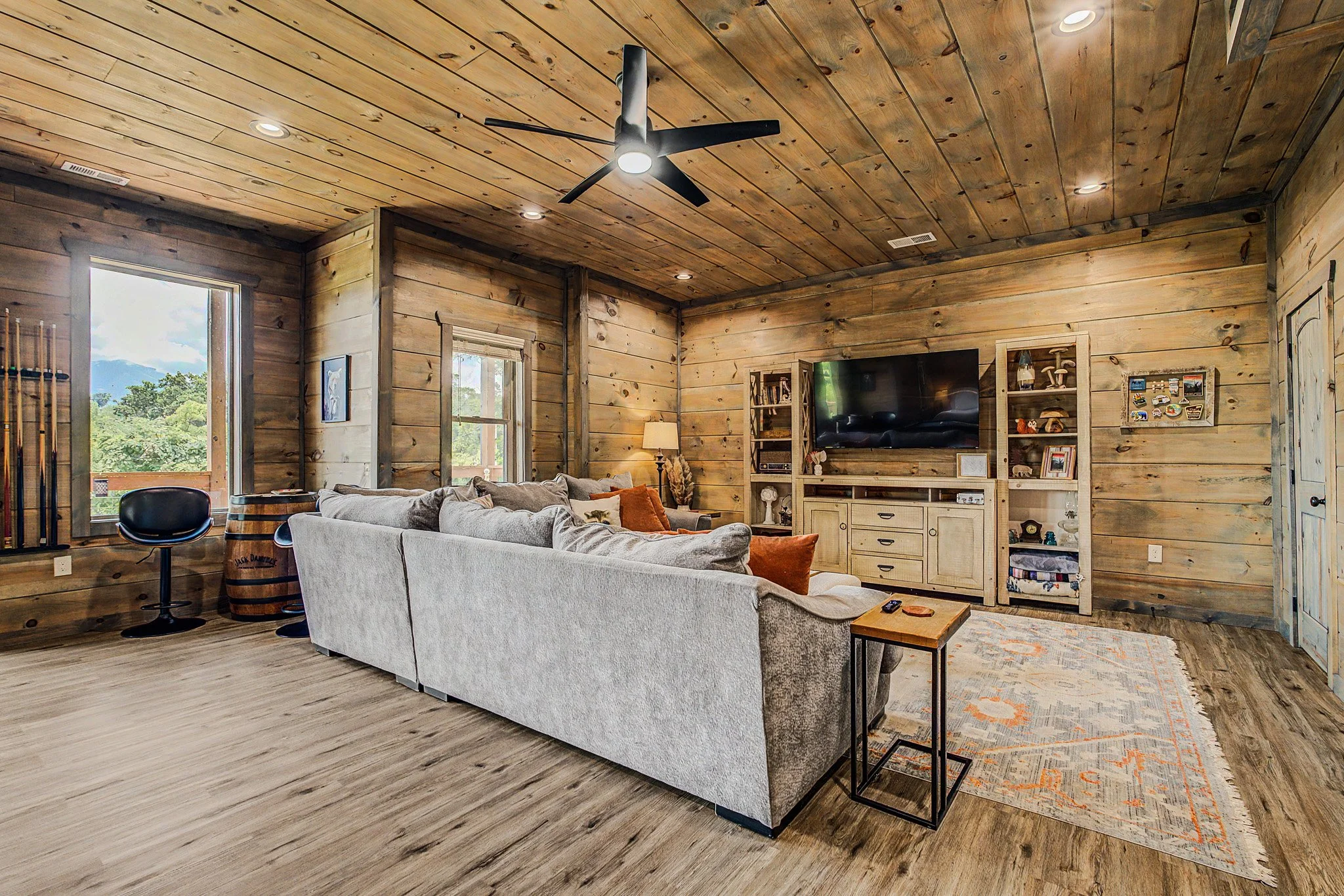 Living room with wood-paneled walls and ceiling, beige sofa, black swivel chair, TV on a wooden cabinet, window revealing a scenic outdoor view, and a patterned area rug.