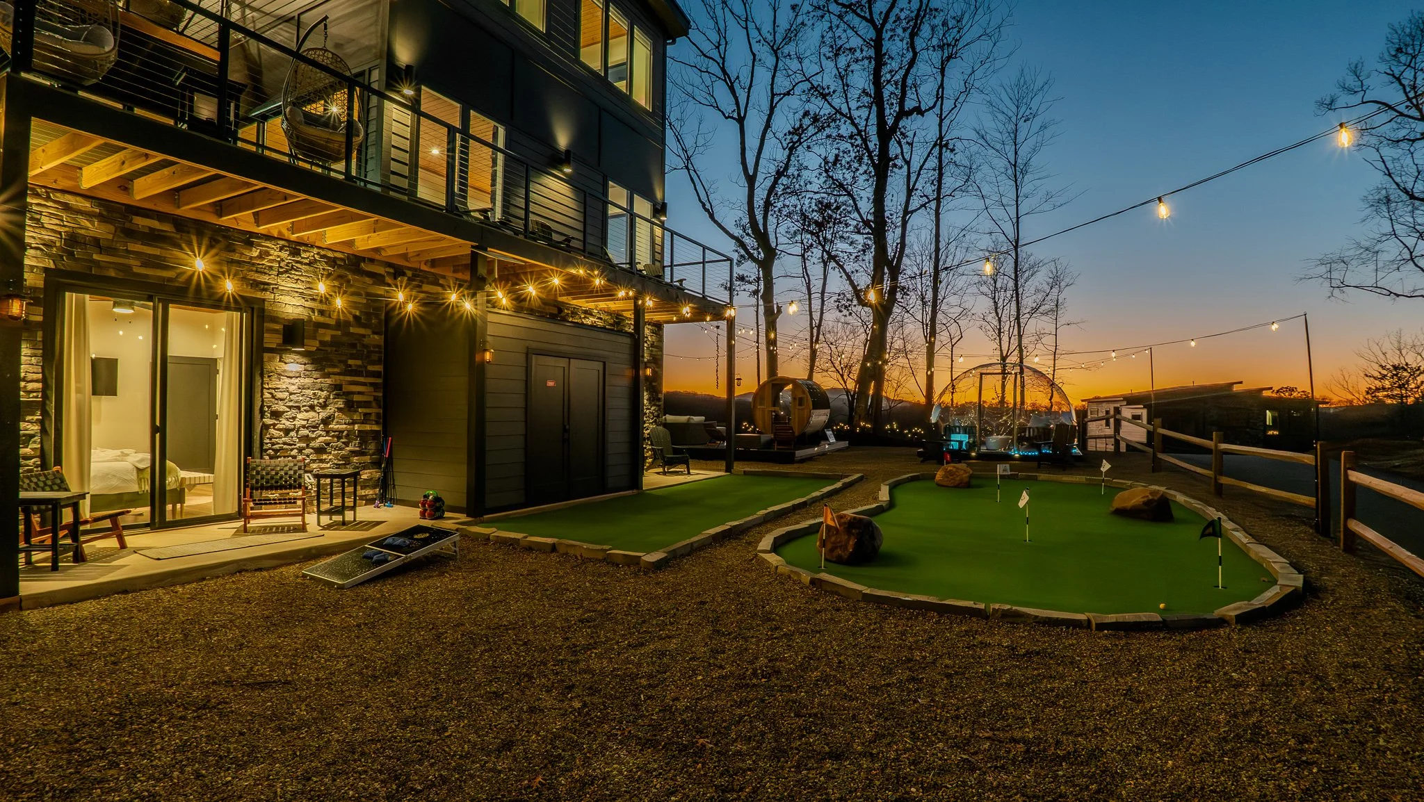 A modern house with a balcony and patio lights at dusk, featuring a mini golf putting green, outdoor seating, and trees with string lights