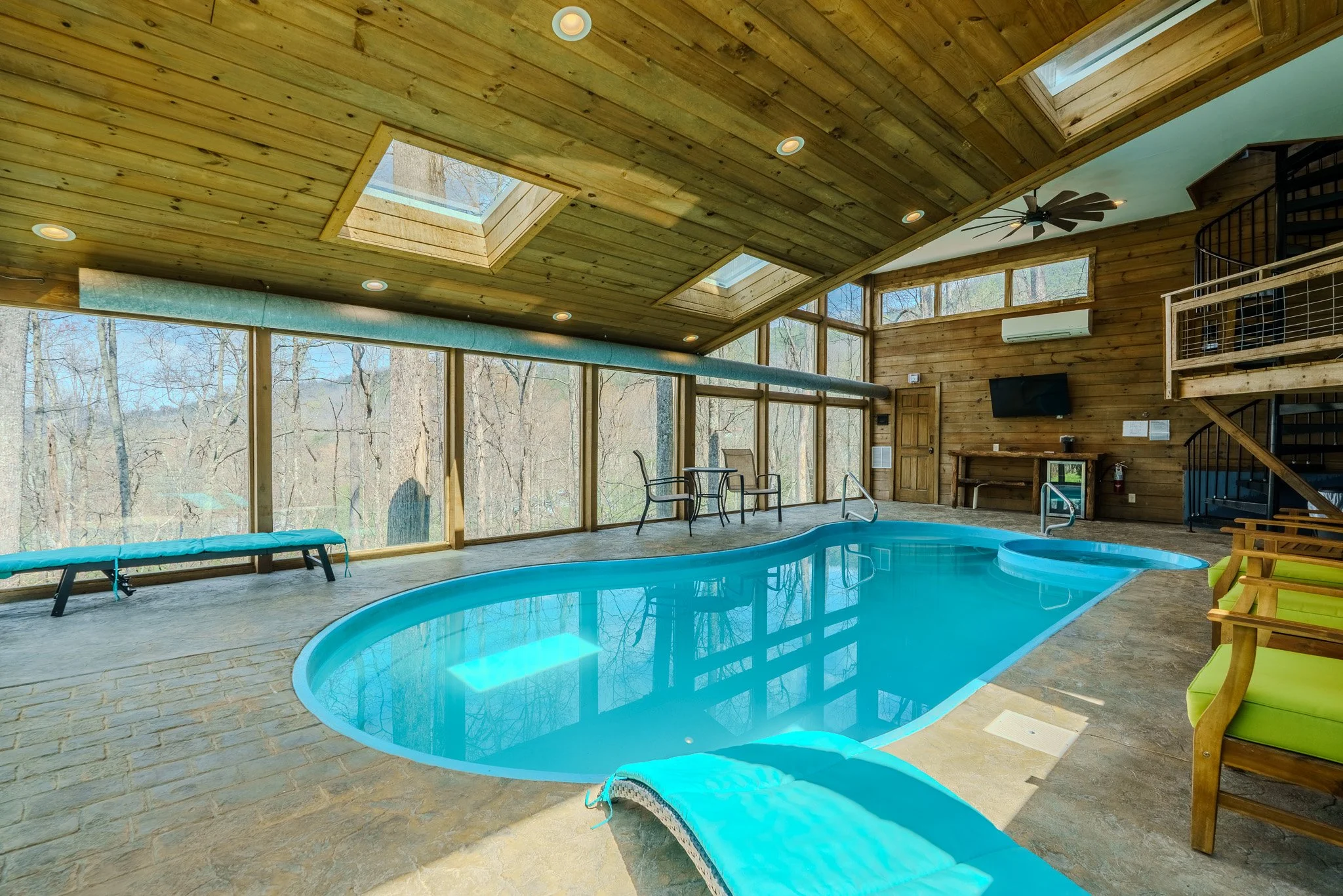 Indoor swimming pool area with large glass windows showing trees outside, wooden ceiling with skylights, poolside chairs, TV, and a spiral staircase.