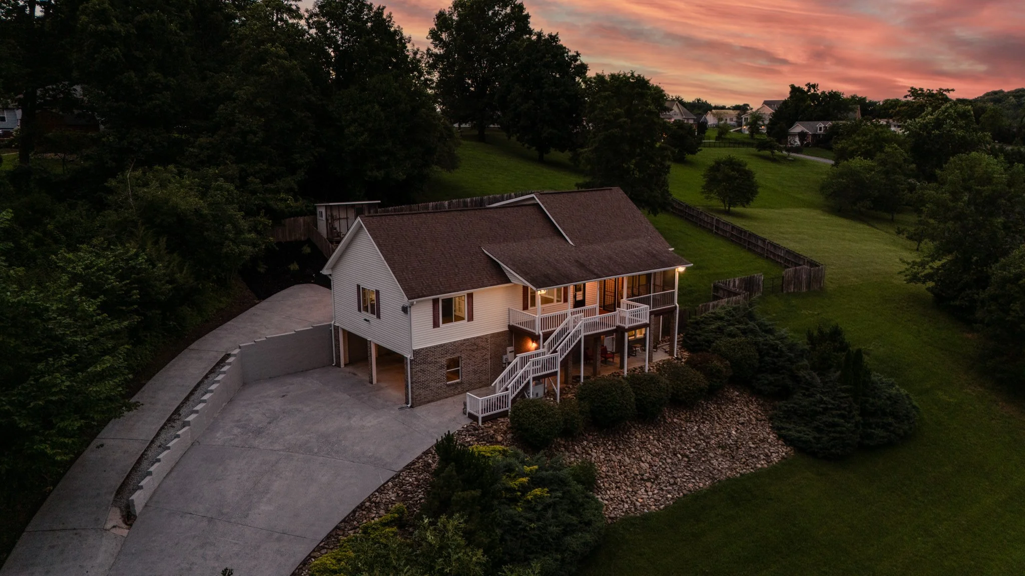 A two-story house with white siding, brick foundation, and a brown shingled roof, illuminated by outdoor lighting at sunset, surrounded by a landscaped yard with trees and an open grassy area, and a curved driveway leading to the house.