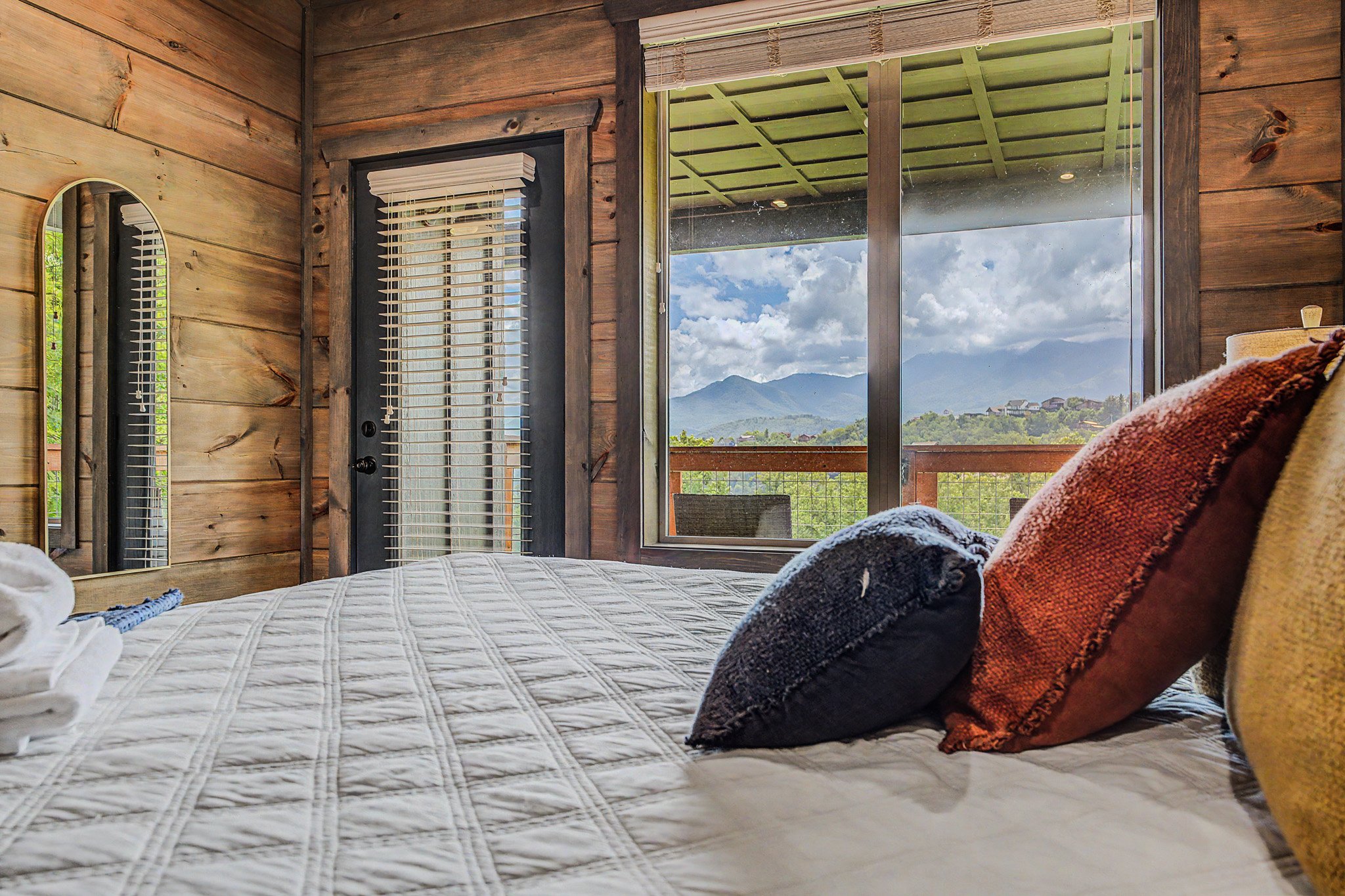 A cozy bedroom with wooden walls, featuring a bed with various colored pillows, a window showing mountains and clouds, and a door with white blinds.