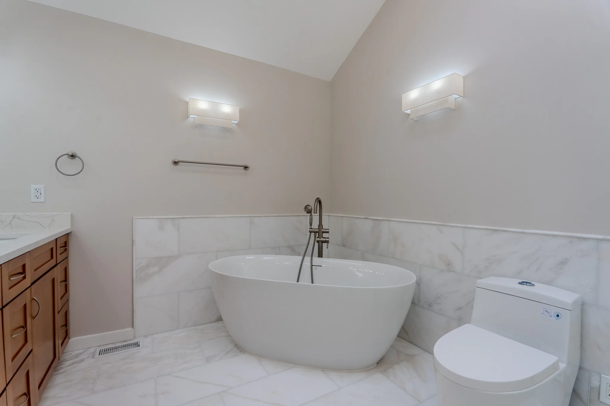 Modern bathroom with a white freestanding bathtub, a toilet, and a vanity with wooden drawers. The walls are painted cream with wall-mounted lights, and there's a marble tile backsplash and floor.
