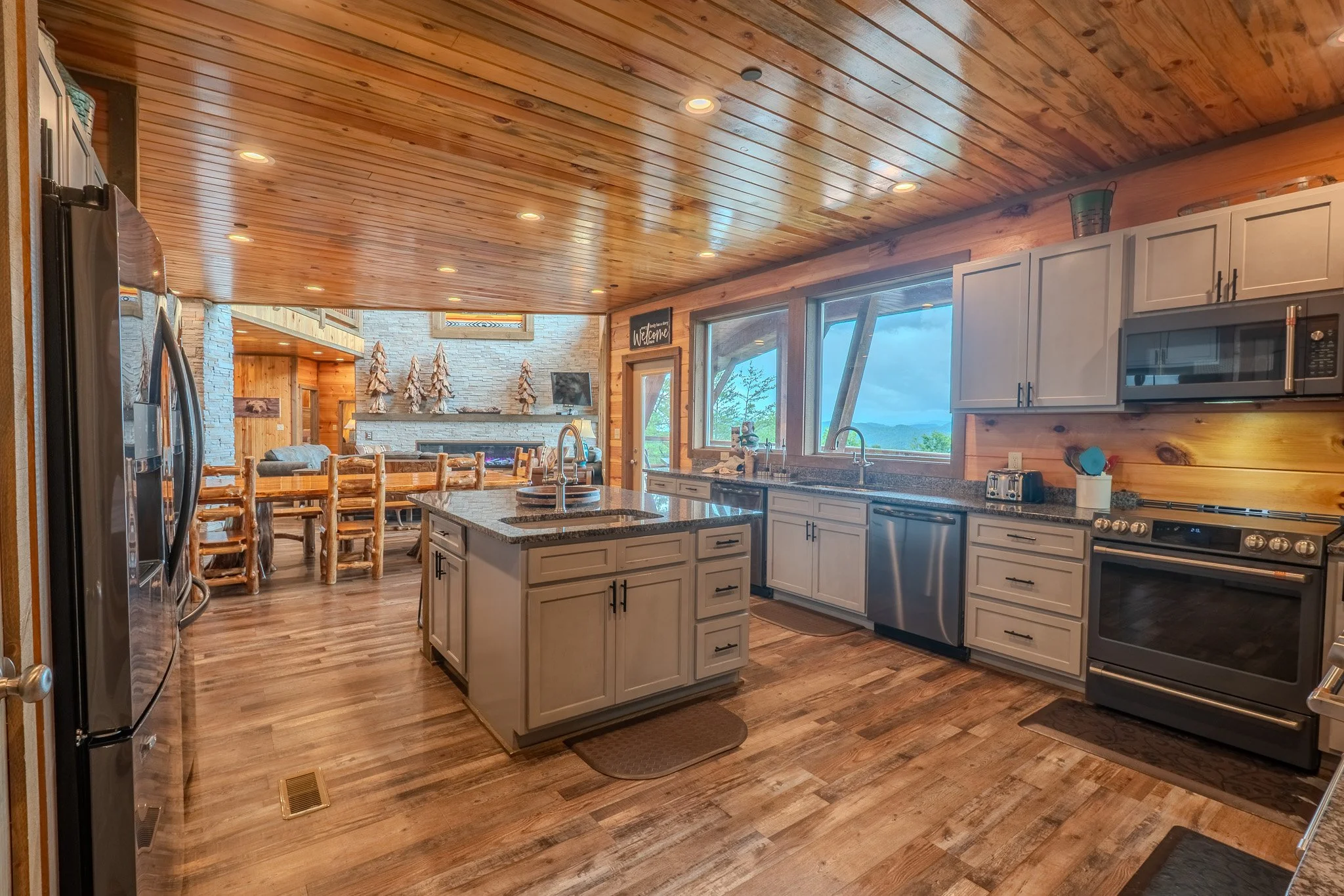 Rustic kitchen with wooden ceiling and floors, white cabinets, stainless steel appliances, large window with view of outdoors, dining table in the background with wooden chairs, decorative trees on mantel, and a fireplace.