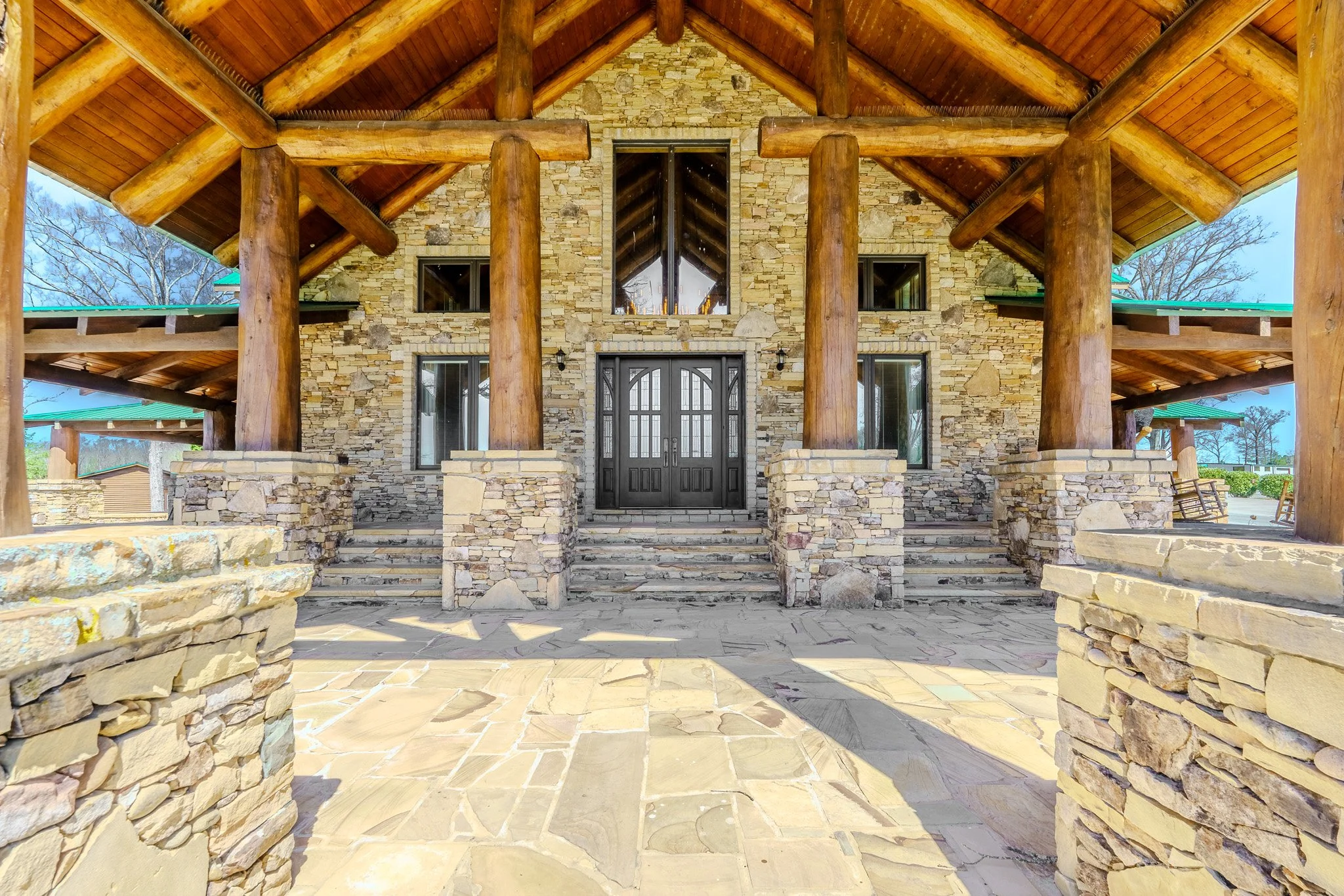 Front view of a rustic stone house with large log columns and a double door entrance, featuring a stone porch and steps.