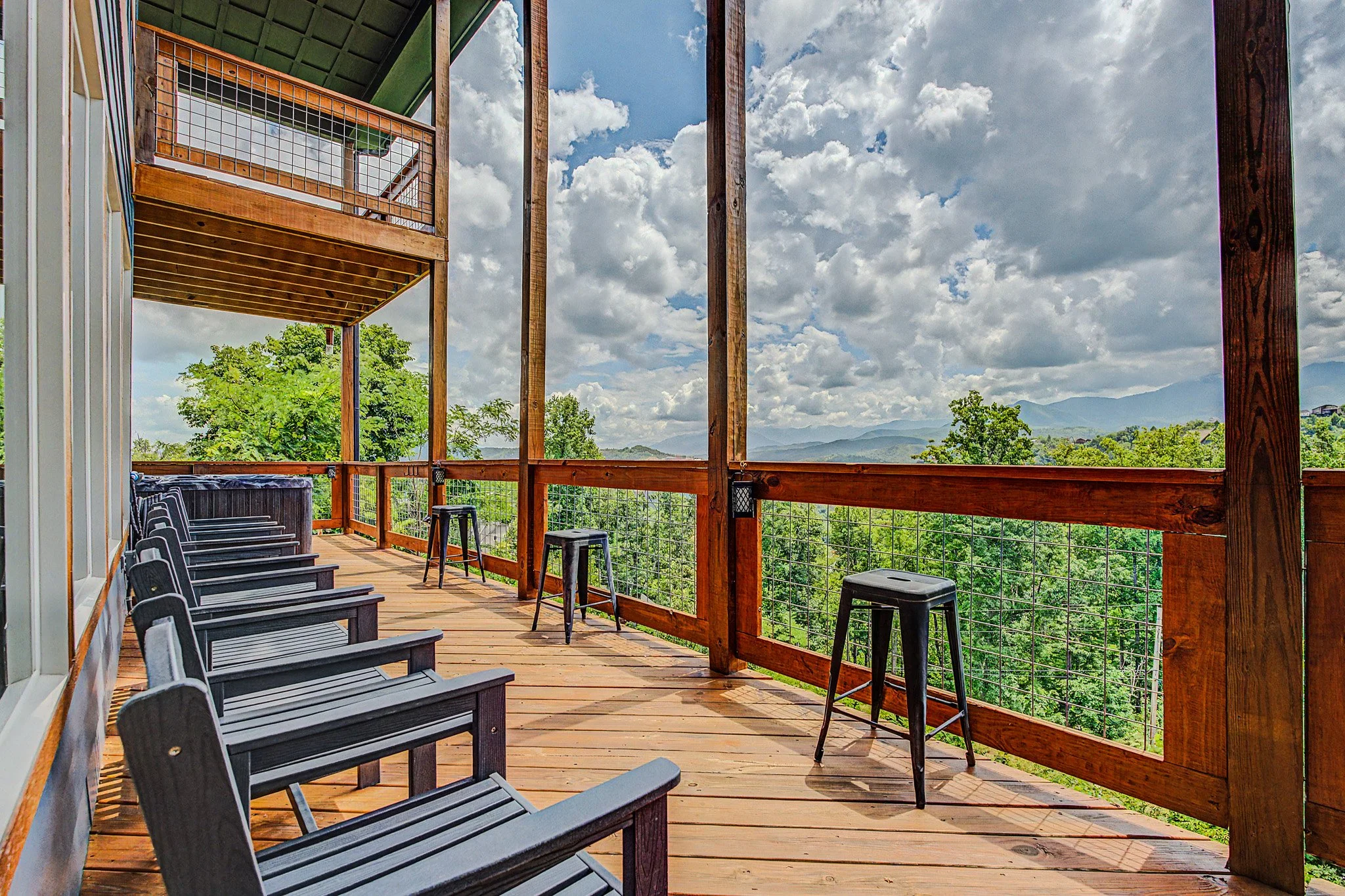 A spacious wooden balcony with black chairs and tall stools, overlooking lush green trees and distant mountains under a partly cloudy sky.