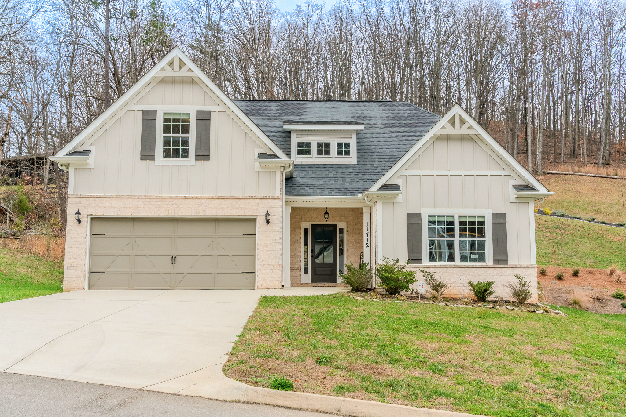 A two-story house with beige siding, black shutters, and a gray shingle roof; front yard with green grass and small bushes; driveway leading to a garage; trees in the background.
