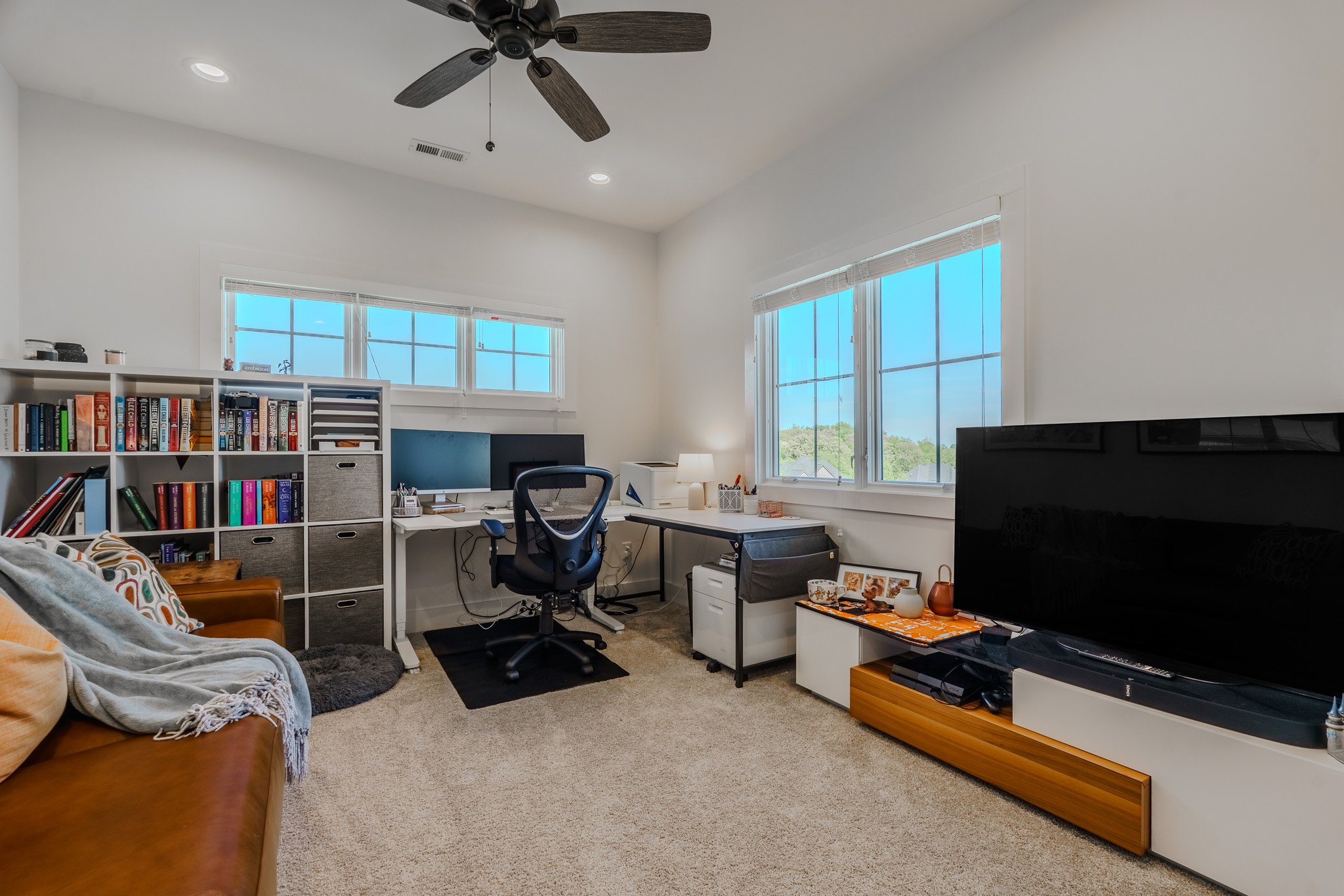 A home office with a white desk, black office chair, bookshelf, TV, and large windows showing a daytime outdoor scene.