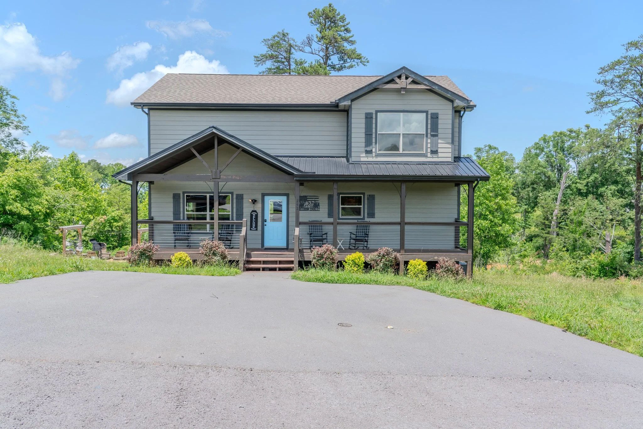 A two-story house with gray siding, a blue front door, a porch with chairs, and surrounded by green trees under a blue sky with clouds.