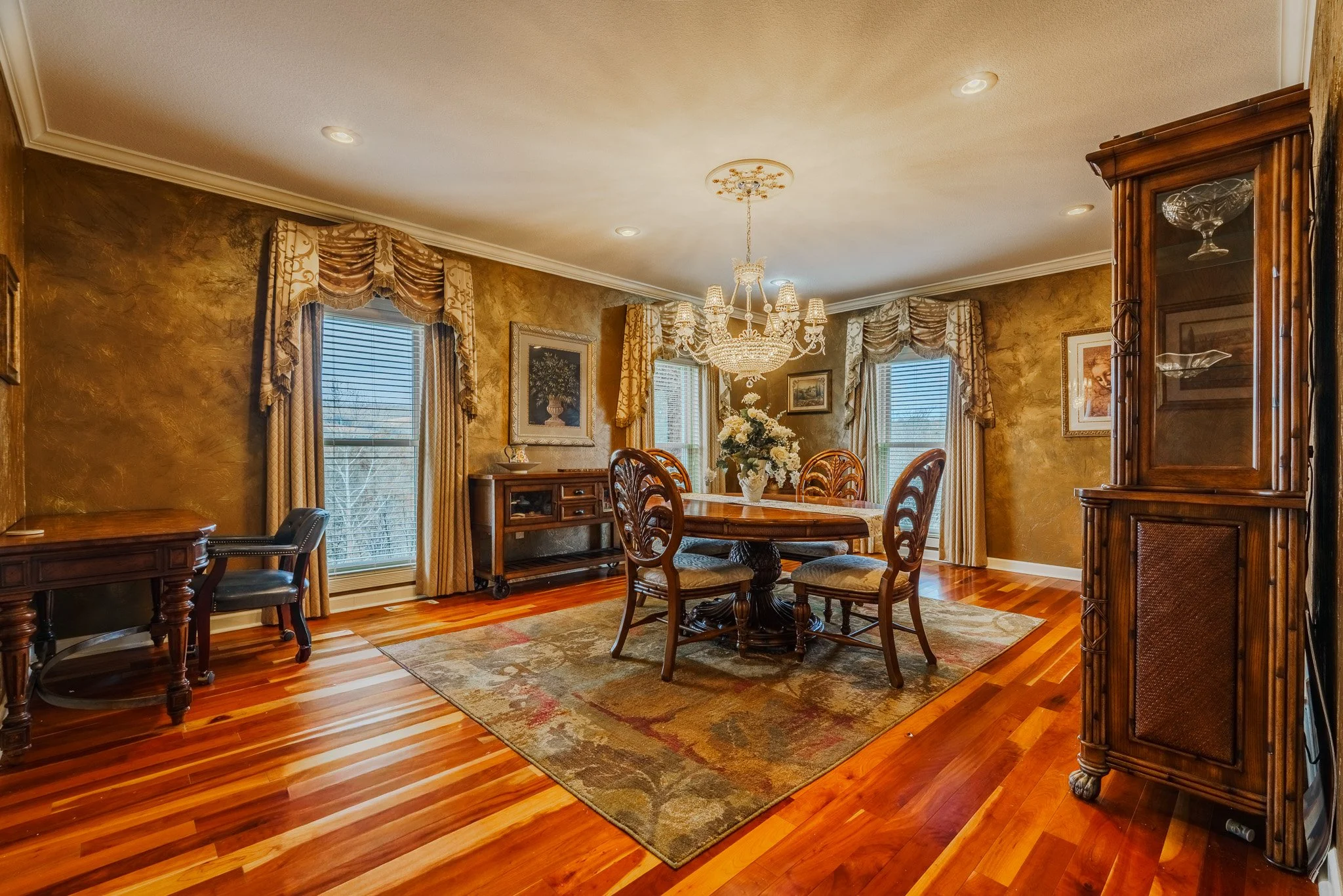 Elegant dining room with a round wooden table, four chairs, a chandelier, floral arrangements, and richly textured gold and cream curtains.
