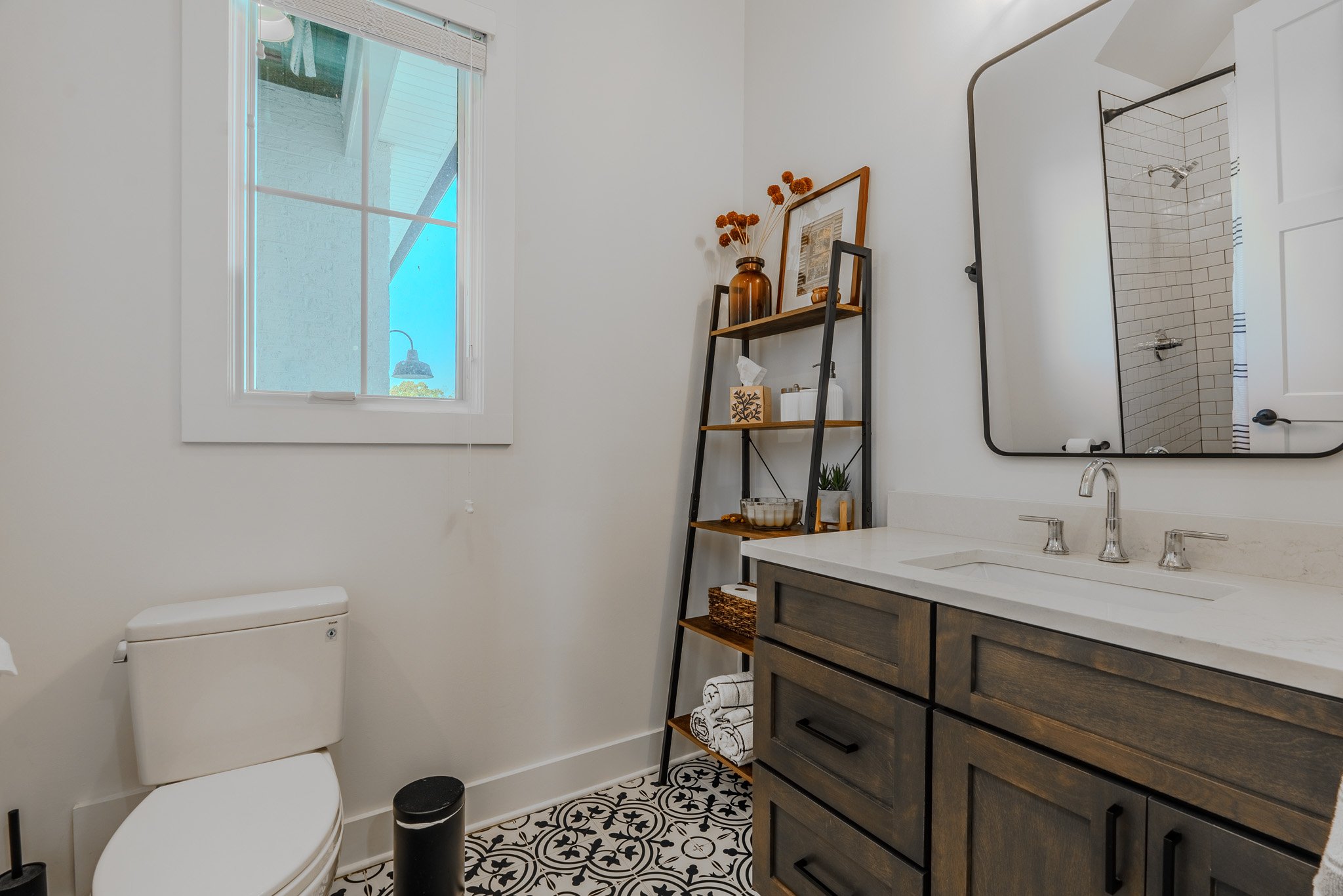 Bathroom with a white toilet, black and white patterned floor tiles, a wooden vanity with a sink, a mirror, and a black ladder shelf. The shelf holds decorative items and towels. A window shows a blue sky and outdoor greenery.