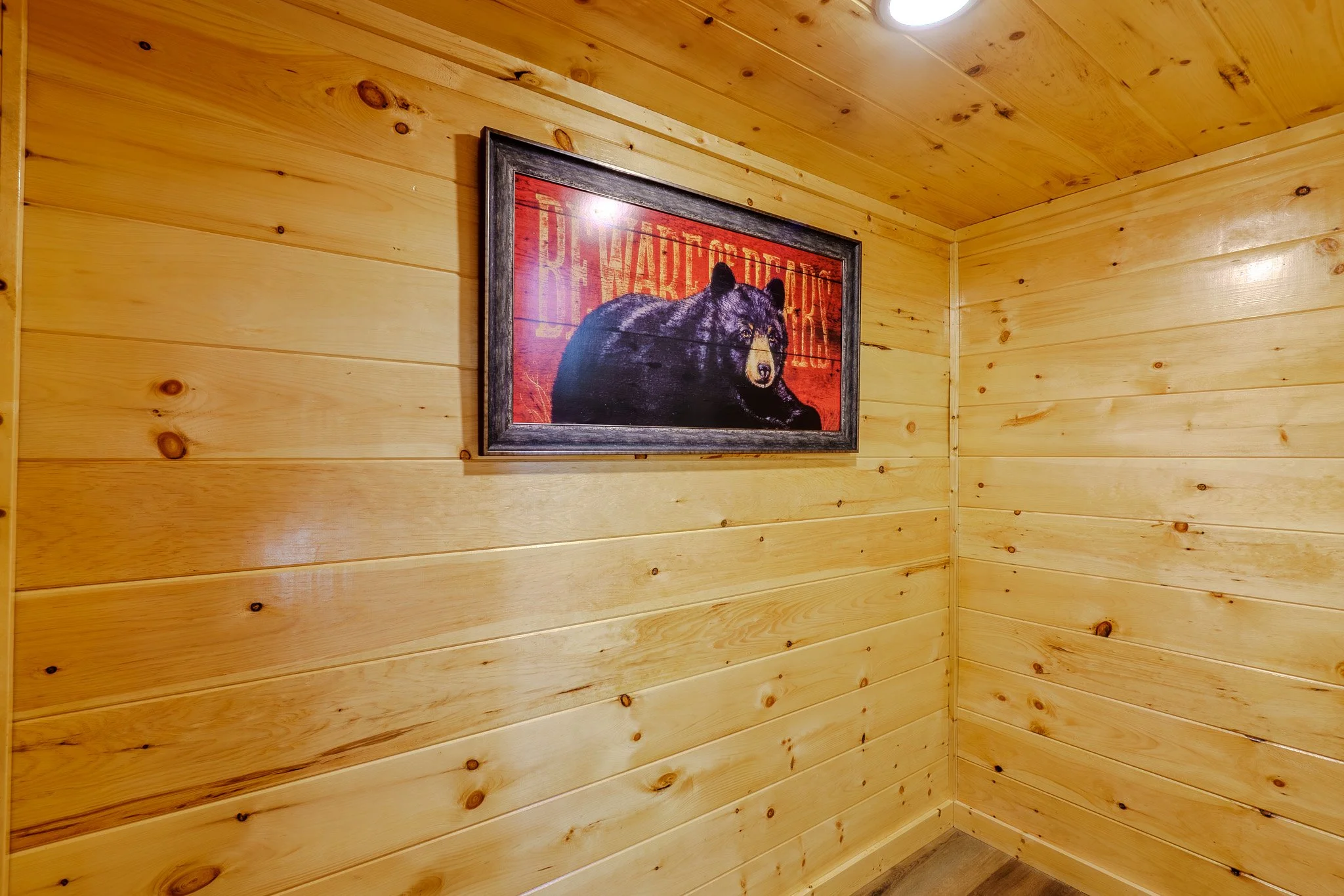 A framed picture of a bear on a red background hanging on a wood-paneled wall in a room with wooden walls and ceiling.