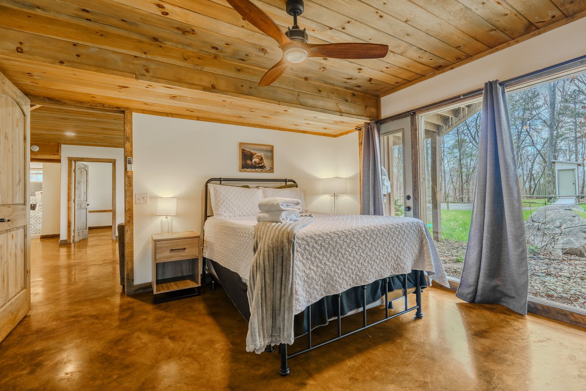 A cozy bedroom with a bed covered in white quilt and folded towels on top, two nightstands with lamps, a ceiling fan, wood-paneled ceiling, and large windows with curtains showing an outdoor view of trees and rocks.