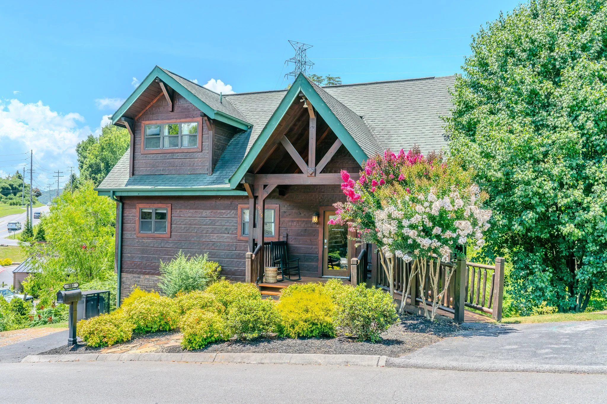 A two-story wooden house with dark brown siding, green trim, and a front porch with black rocking chairs amid landscaped bushes and flowering plants, on a sunny day with blue sky.