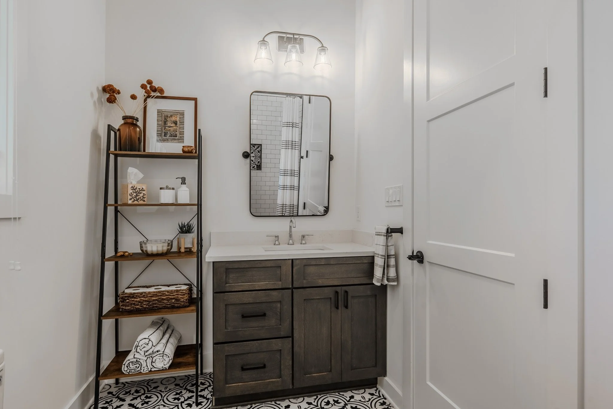 A modern bathroom with a dark wood vanity, white countertop, and a rectangular mirror above the sink. There is a black metal and wood shelving unit to the left with decorative items and towels. The bathroom also has black and white patterned floor ti