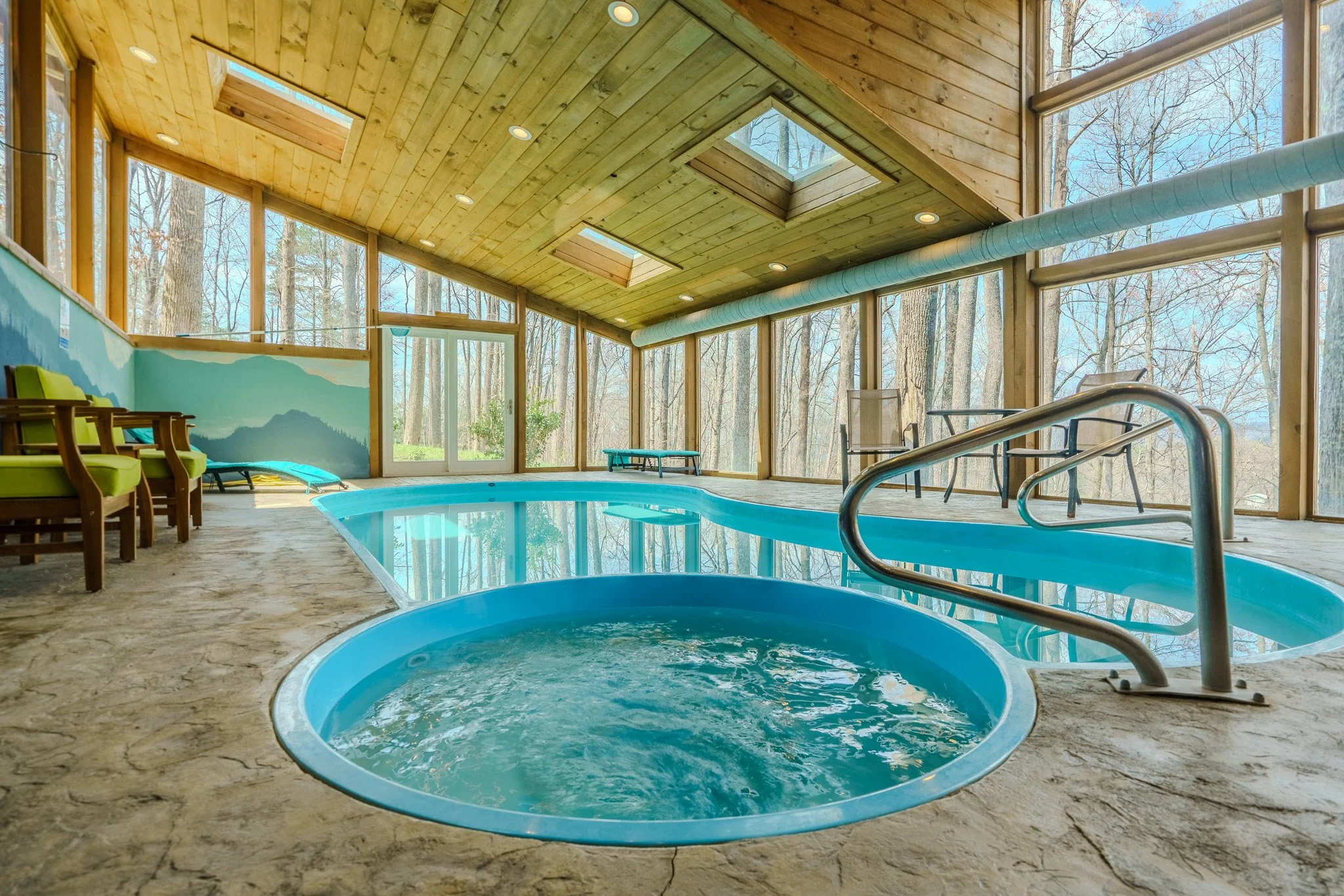 Indoor pool area with a small circular hot tub, surrounded by chairs, large windows showing trees, skylights, and wooden ceiling.