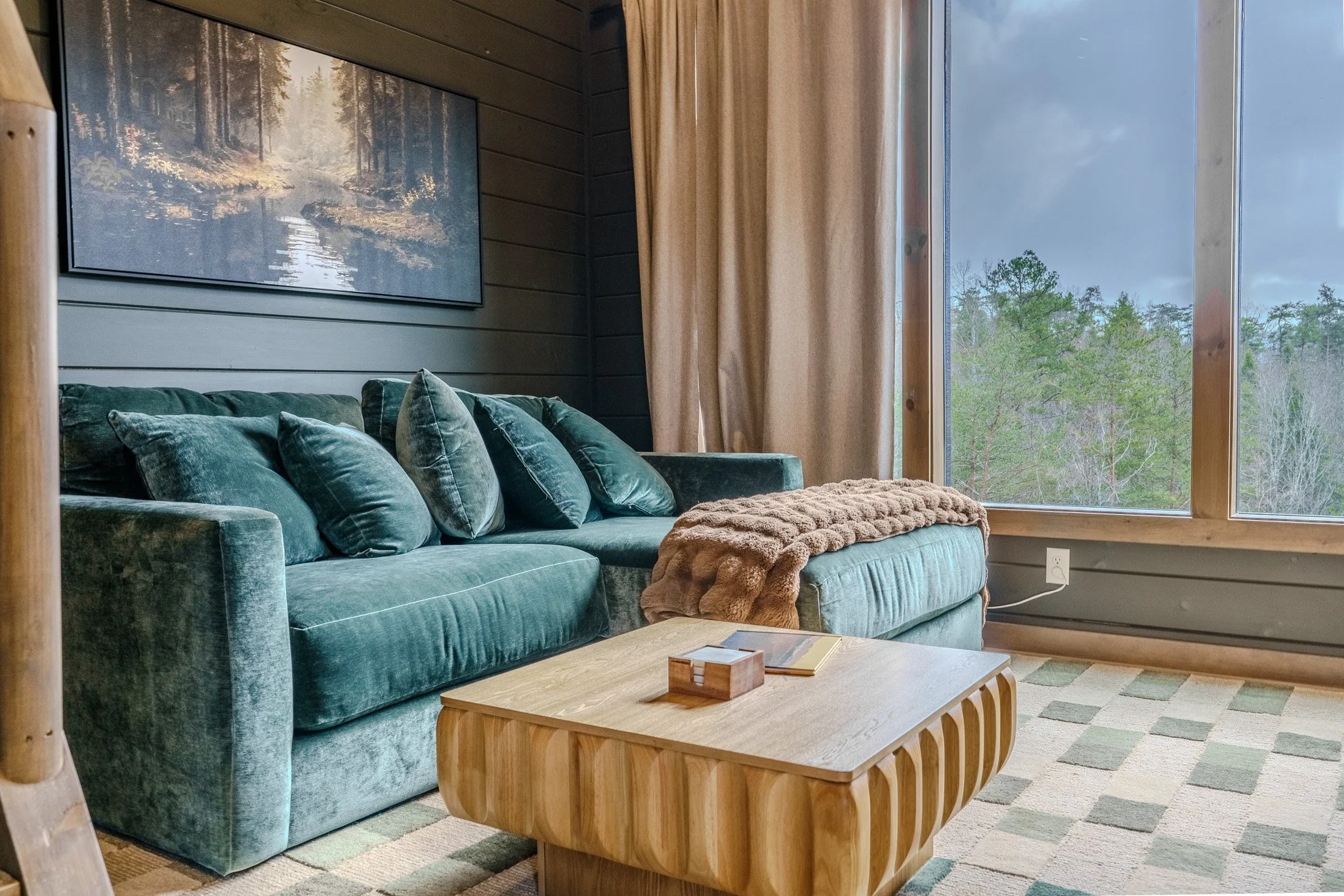 Living room with a green velvet sofa, multiple matching pillows, a wooden coffee table, a window showing trees outside, and a brown furry blanket on the sofa.