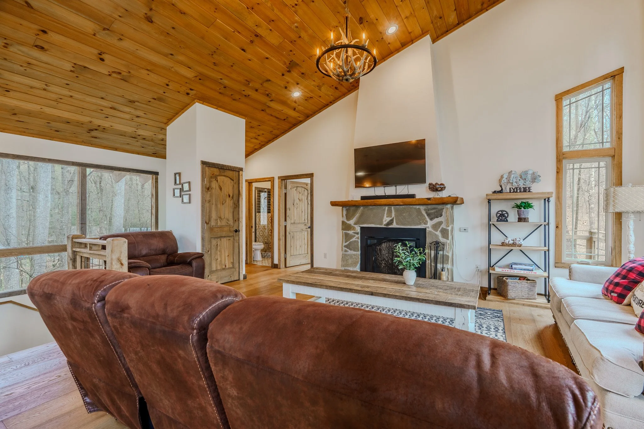 Living room with wooden ceiling, leather and fabric sofas, stone fireplace, mounted TV, and large windows showing trees outside