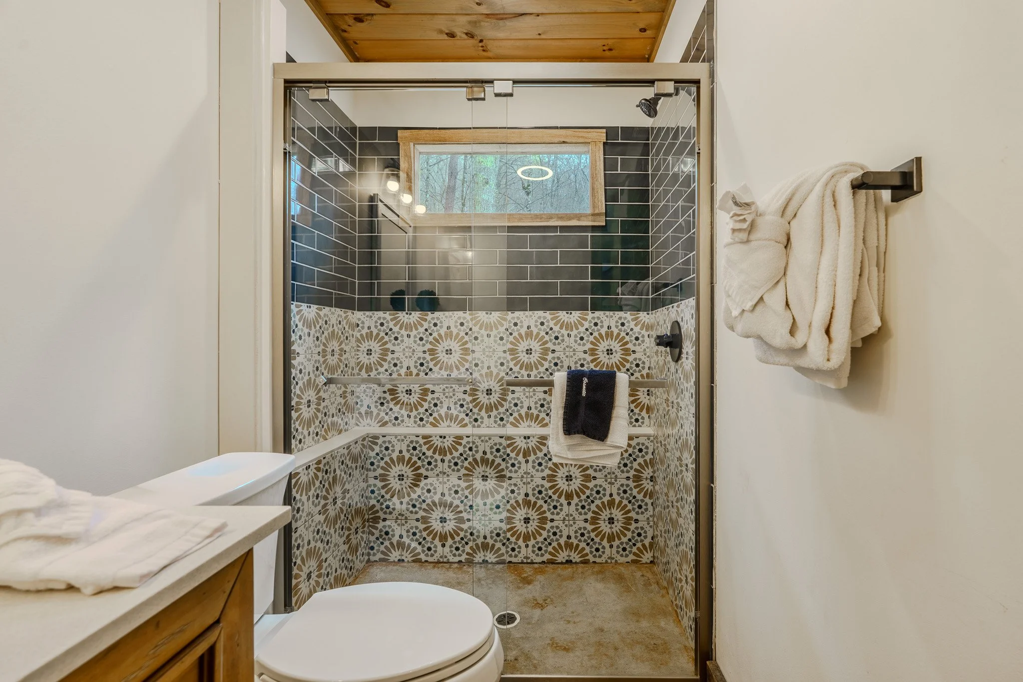 A small bathroom with a shower enclosed by glass, with patterned tile walls and a window, alongside a white toilet and towel racks.