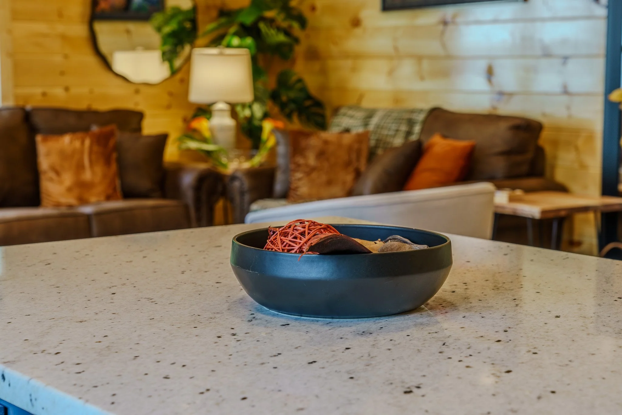 A black bowl with decorative red, black, and gray objects on a white speckled countertop in a cozy living room with wooden panel walls, brown and beige sofas, and warm lighting.