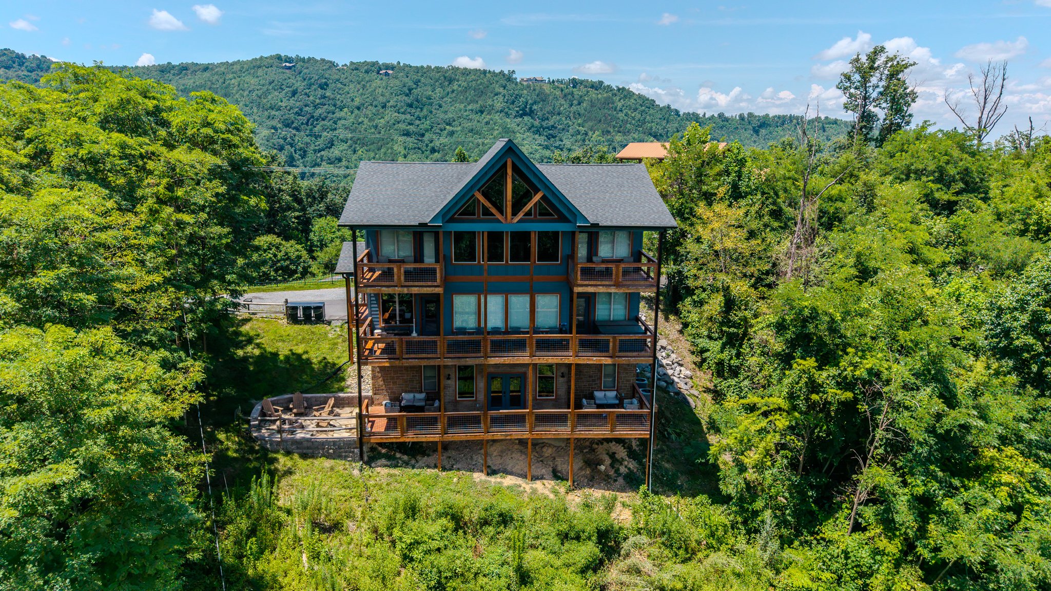 A multi-story house surrounded by lush green trees, with a mountain range and a partly cloudy sky in the background.