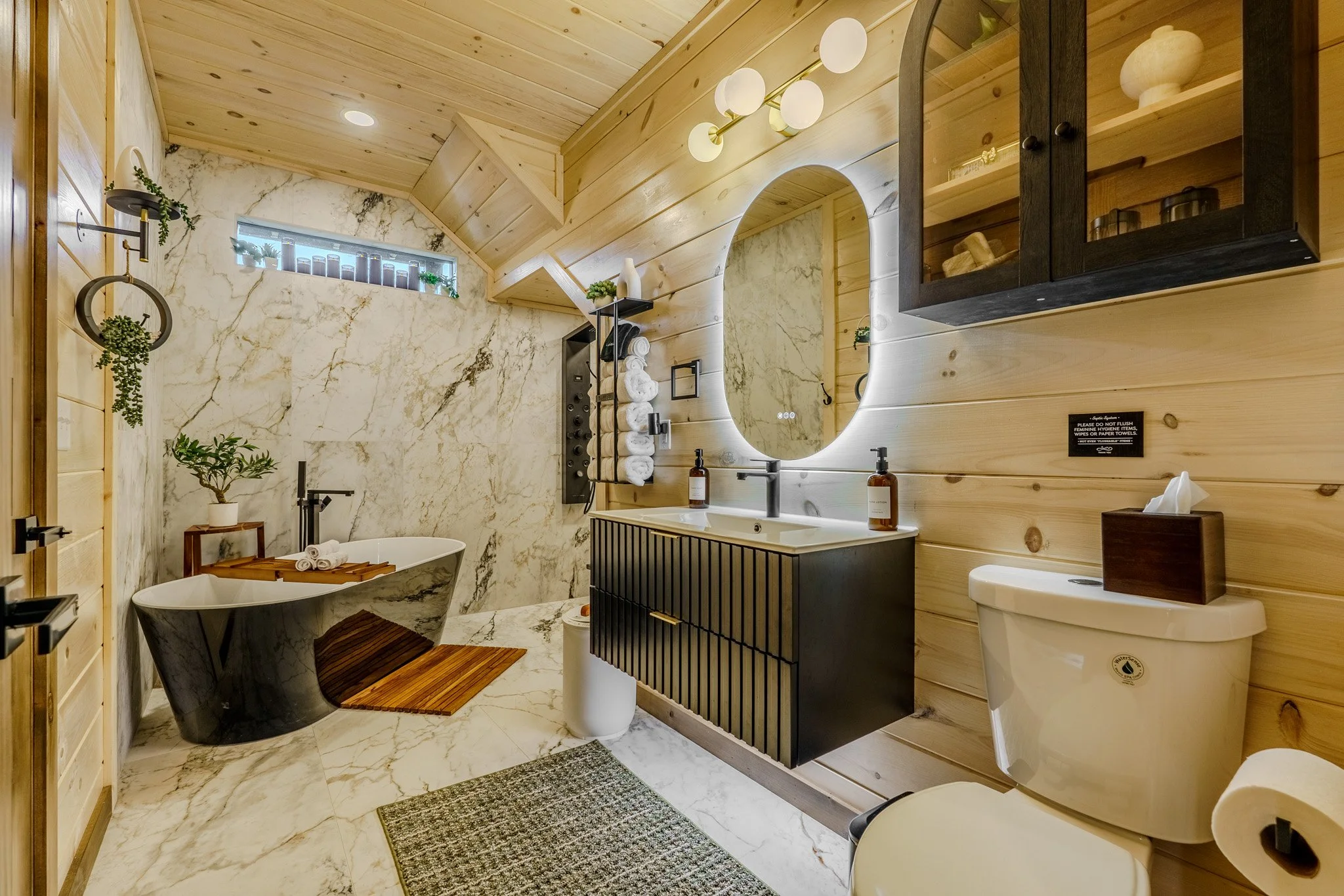 Modern bathroom with wood-paneled walls and marble floor and shower area. It features a black bathtub, a black vanity with a white sink, a large backlit mirror, and a toilet. Decorative plants and wooden accents are also present.