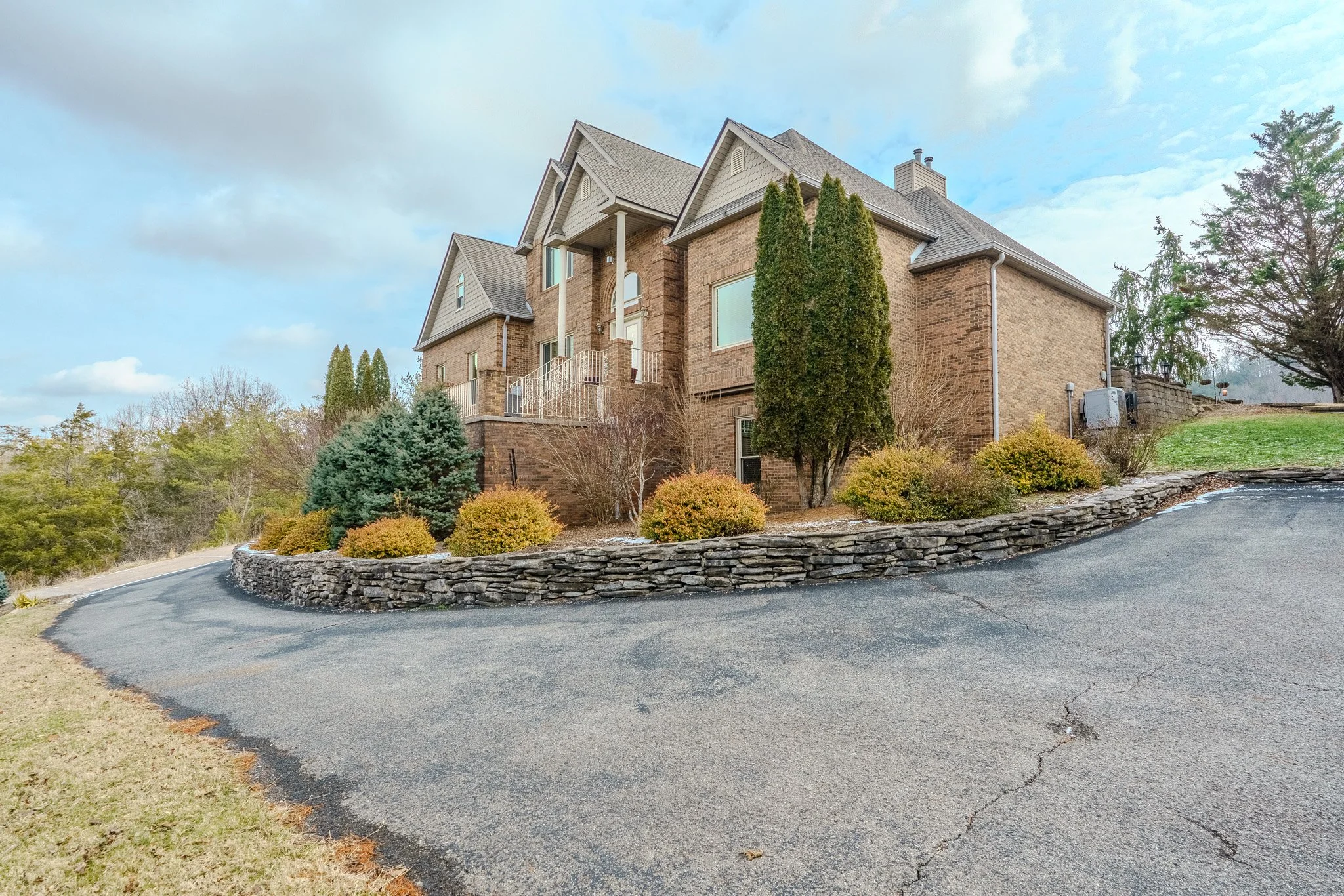 A large brick house with multiple gabled roofs, large windows, and a front porch, surrounded by landscaped bushes and trees, on a slightly elevated terrain, viewed from the curved driveway.