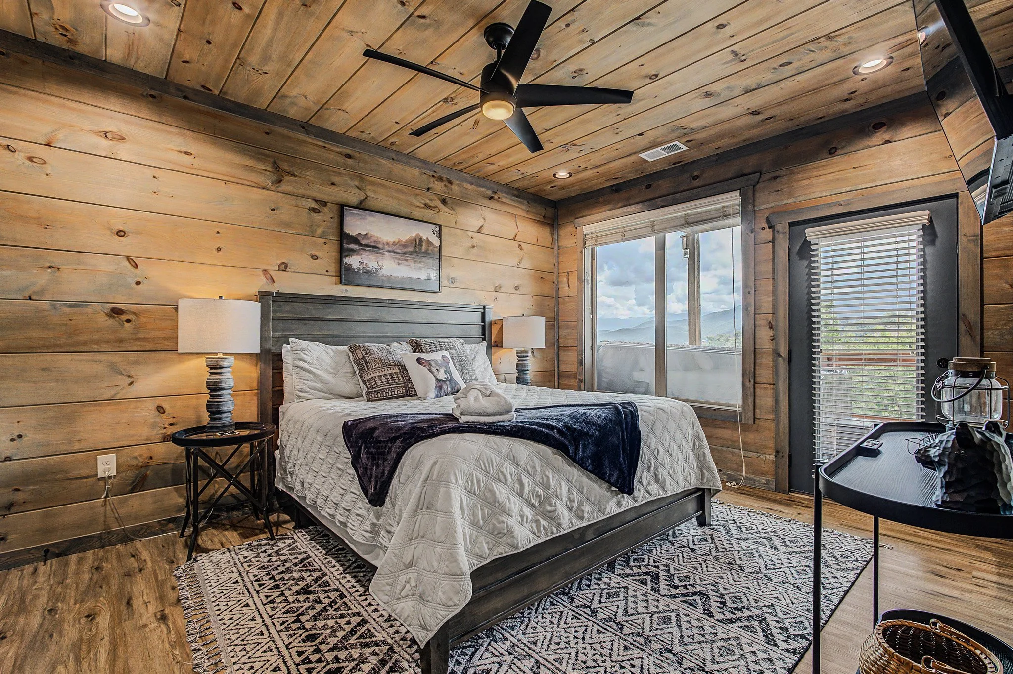A bedroom with wooden walls and ceiling, a large bed with white bedding and decorative pillows, two nightstands with lamps, and a rug. There are windows showing an outdoor view, and a television mounted on the wall.
