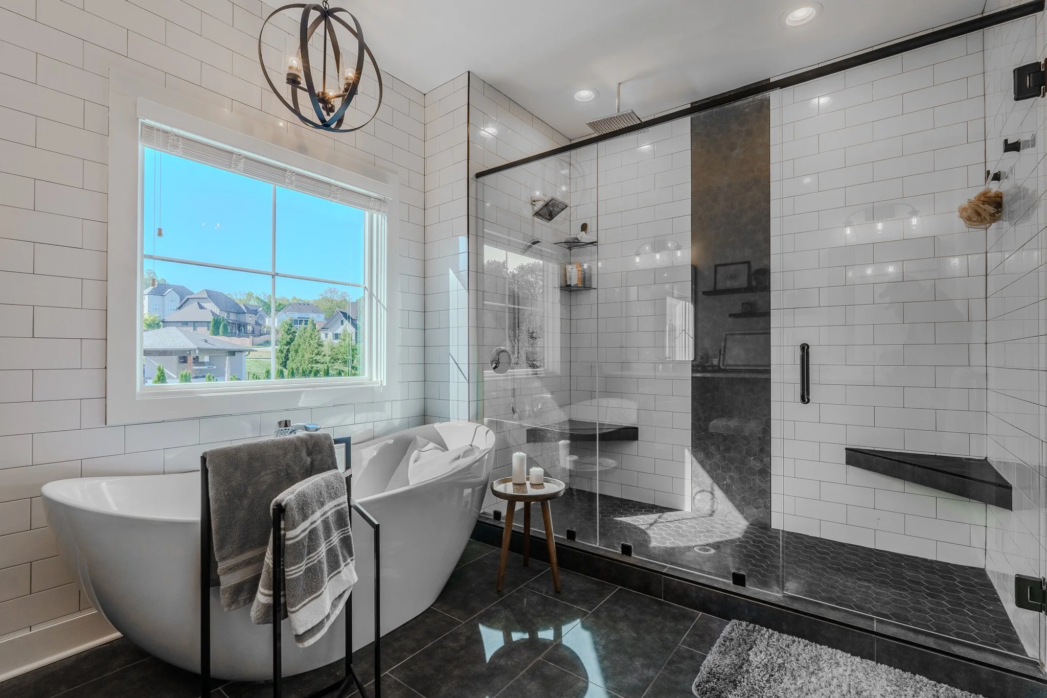Modern bathroom with white subway tile walls, a large window, a black-framed glass walk-in shower, a freestanding bathtub with towels, a small wooden stool with candles, and a gray bath mat.