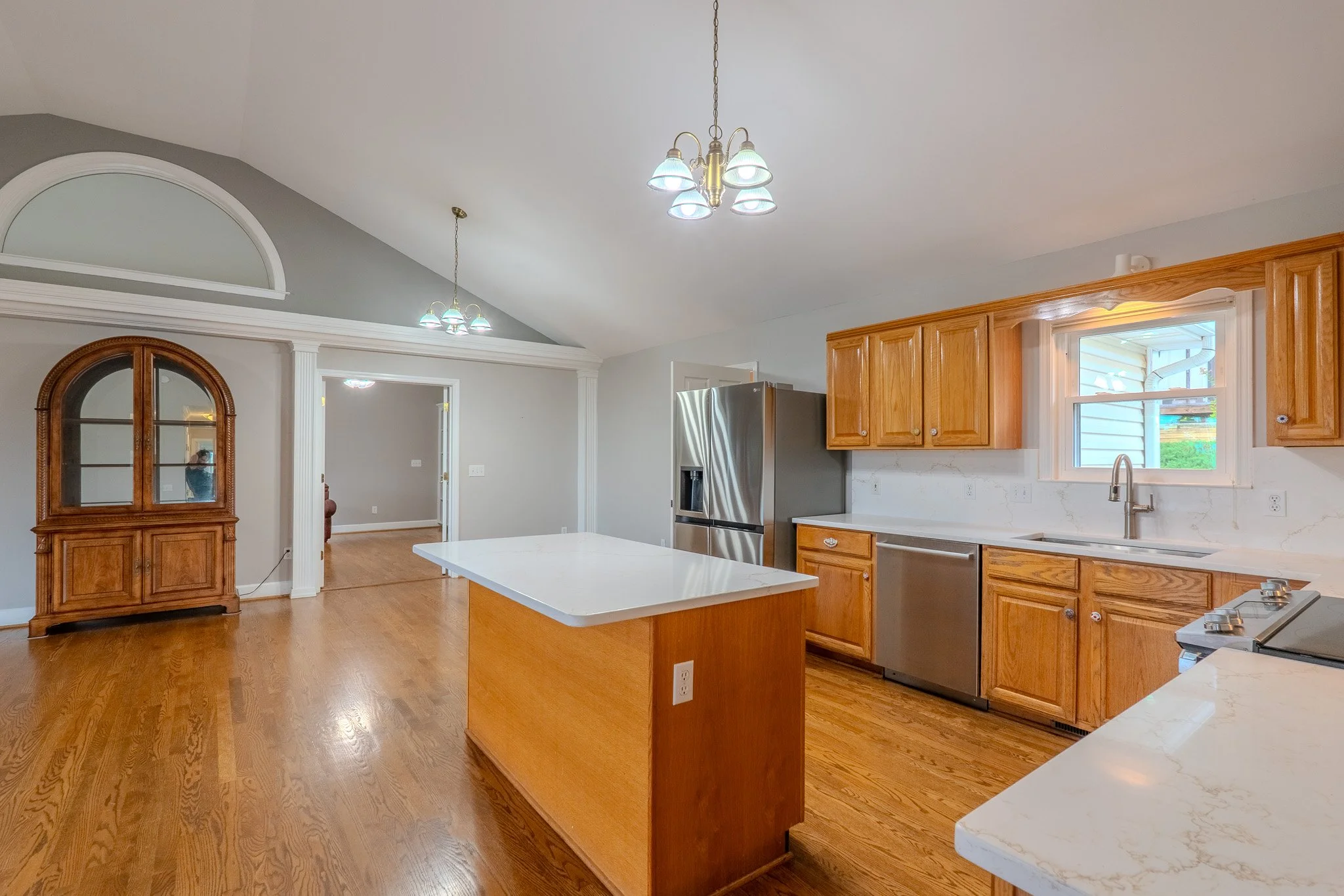 Empty kitchen with wooden cabinets, stainless steel refrigerator, white countertops, and hardwood floors. A kitchen island and a window above the sink are visible.