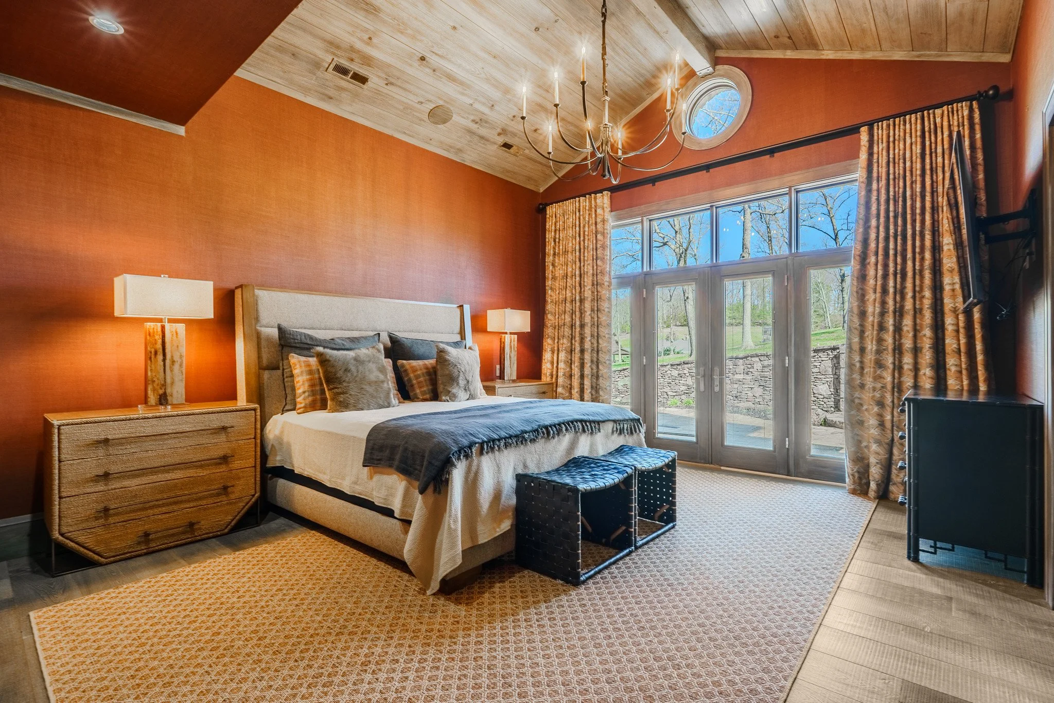 Bedroom with orange textured wall, beige upholstered bed with multiple pillows, wooden nightstands with lamps, large window with patterned curtains, chandelier, flat-screen TV, outdoor view of trees and stone wall, on wood and patterned area rug.