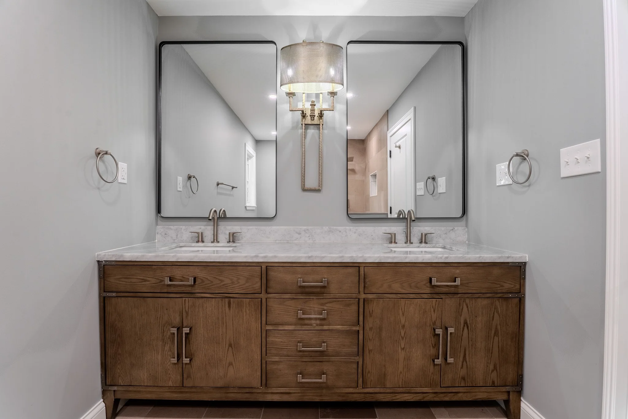 A bathroom vanity with a wood cabinet, marble countertop, two mirrors, and two sinks, with a light fixture in the center.