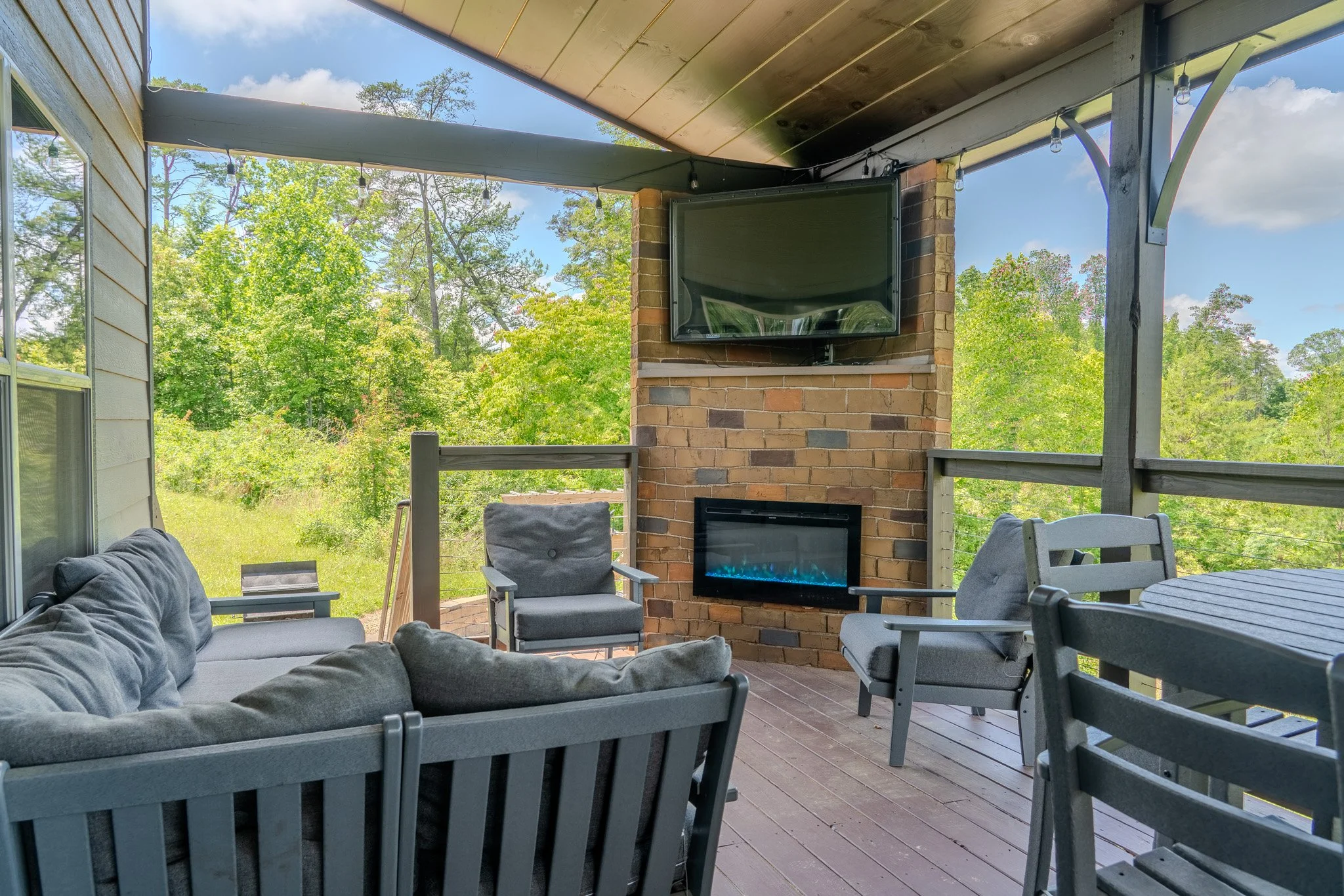 Outdoor covered porch with gray outdoor furniture, a wall-mounted television, and a fireplace, surrounded by green trees and a blue sky.