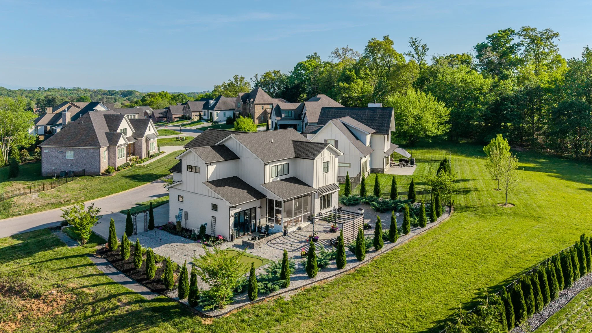 Aerial view of a suburban neighborhood featuring a modern white two-story house with a backyard, surrounded by green grass and trees, with several other houses in the background and a wooded area behind.