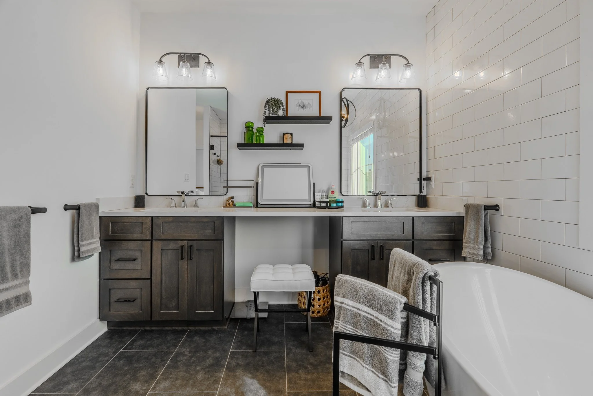 Modern bathroom with a double sink vanity, dark wooden cabinets, large mirrors, and a bathtub on the right. The walls are white with one accented tile wall. Towels hang on black towel bars.