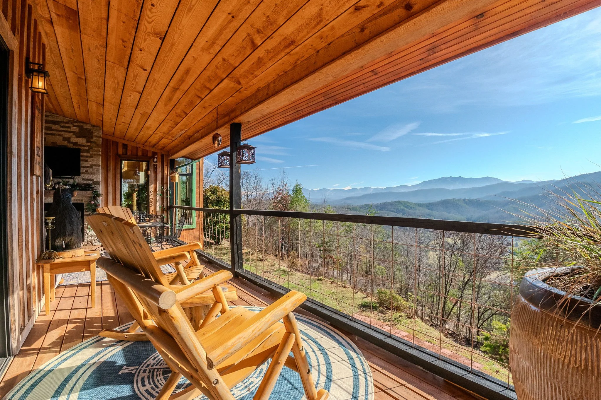 View of a wooden balcony with two wooden rocking chairs, potted plant, and lanterns overlooking a scenic mountain landscape on a clear day.