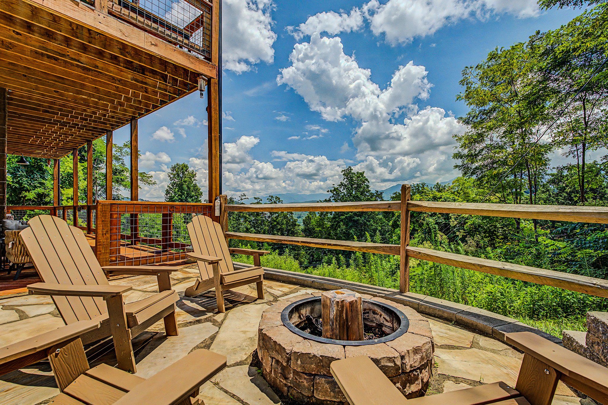 A wooden deck with Adirondack chairs surrounding a fire pit, overlooking a lush green landscape with trees and mountains under a partly cloudy sky.