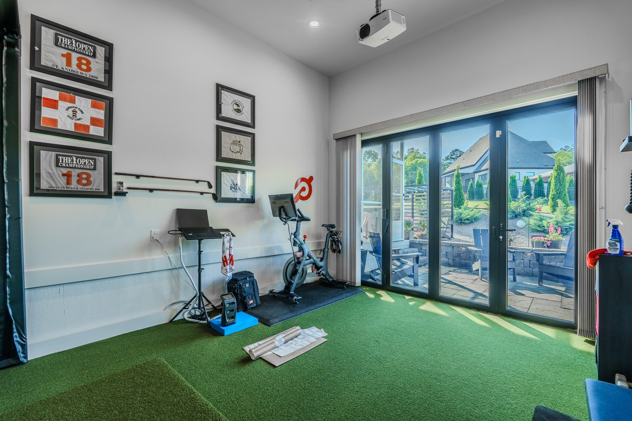 Indoor golf simulation room with sliding glass door leading to patio, framed sports photos on wall, exercise bike, and fitness equipment.