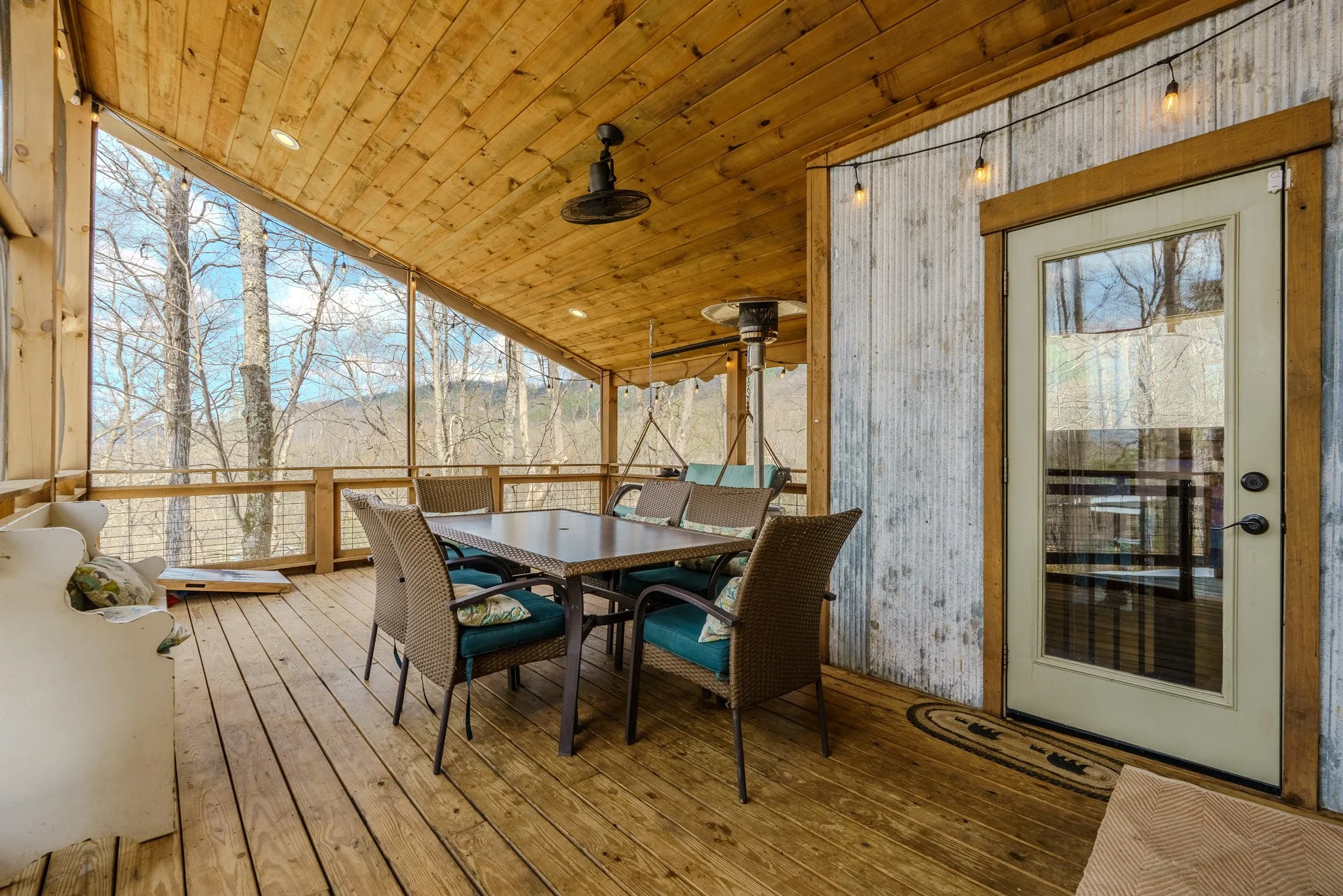 A covered outdoor porch with a wooden ceiling, wooden flooring, and a view of trees in the background. Featuring an outdoor dining table with six chairs, a swing, a ceiling fan, string lights, and a door with a glass pane leading outside.