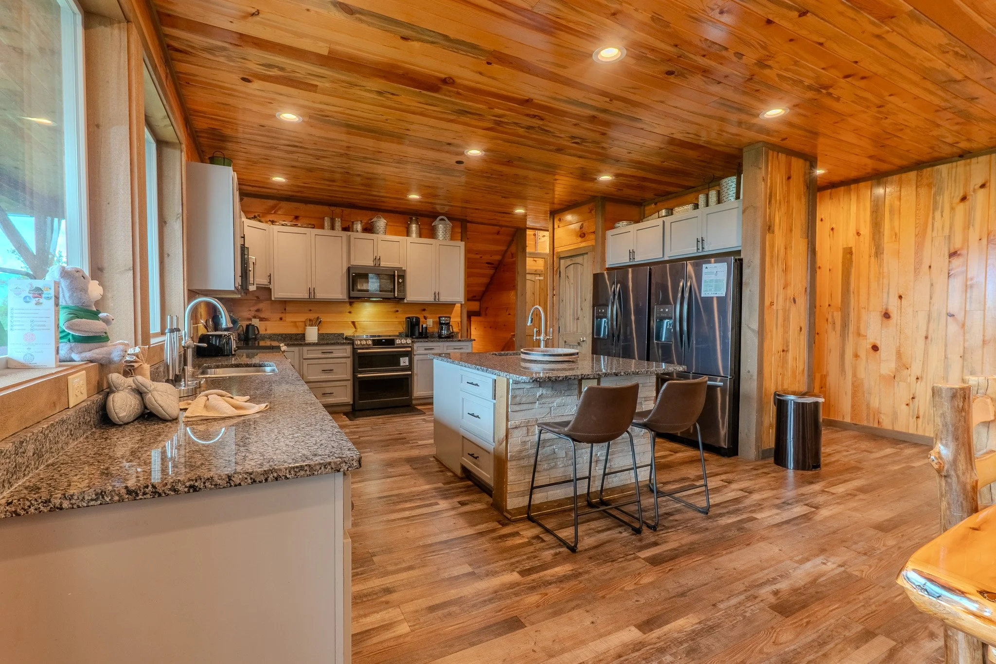 Rustic kitchen with wooden walls and ceiling, granite countertops, stainless steel appliances, an island with bar seating, and wooden flooring.