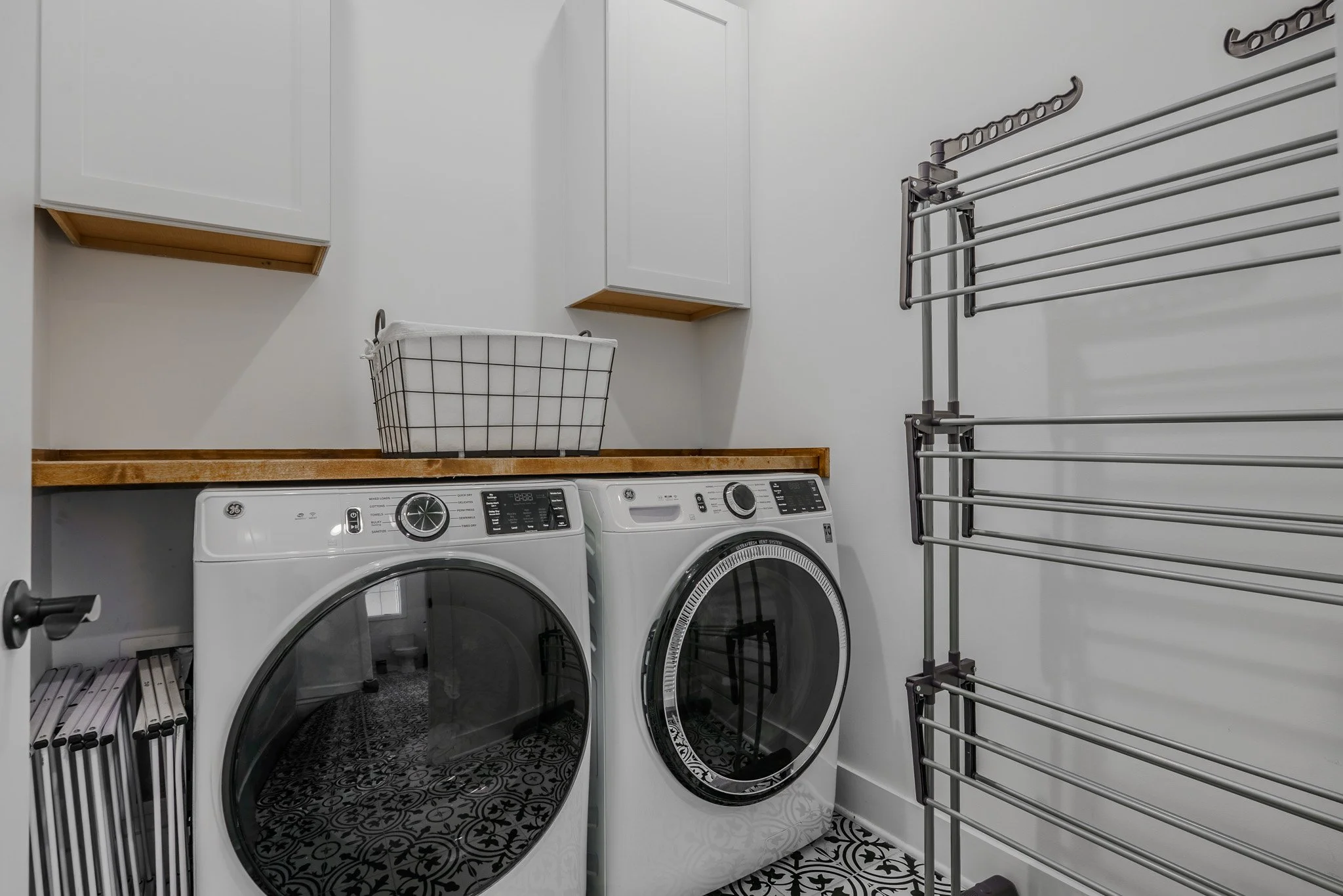 Laundry room with front-loading washer and dryer, white cabinets, a wire laundry basket on a wooden counter, and a metal drying rack on the wall.